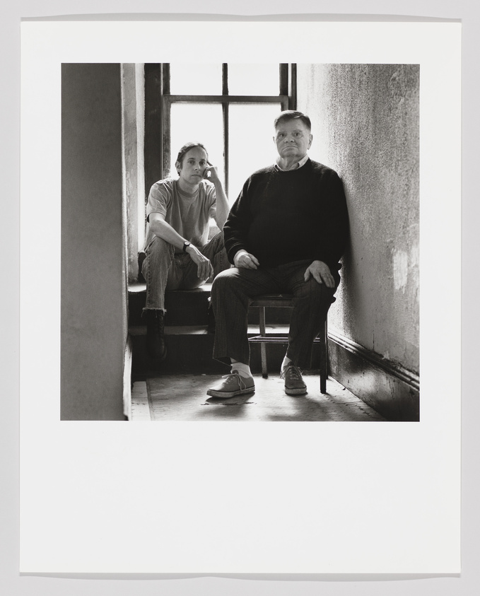 Two people sitting by a window in a dimly lit room, one gazing outside, the other looking at the camera.