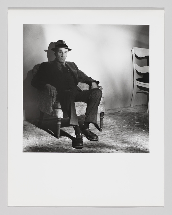 Man in suit and fedora sitting in a chair, with a shadow of a chair on the wall behind him.