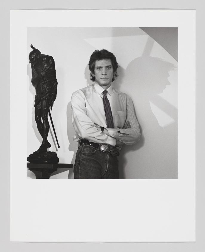 Man in dress shirt and tie with arms crossed, standing beside a dark statue, with a shadow cast on the wall.