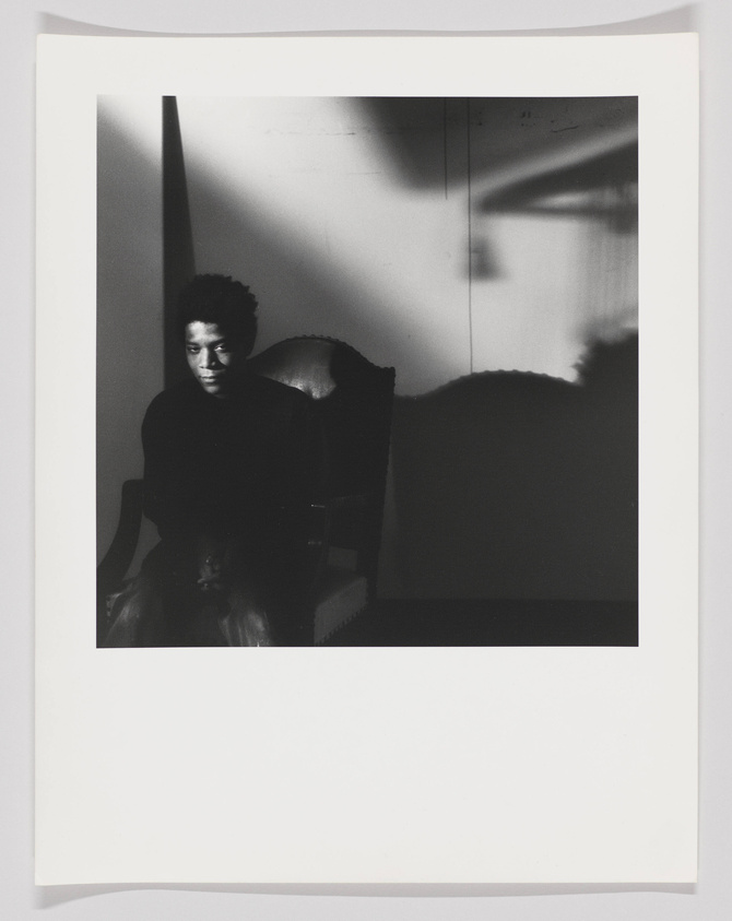 A black and white photograph capturing a person sitting in a dark room with strong light and shadow contrasts. The individual appears contemplative, seated in an ornate chair, with shadows casting patterns on the wall and floor.