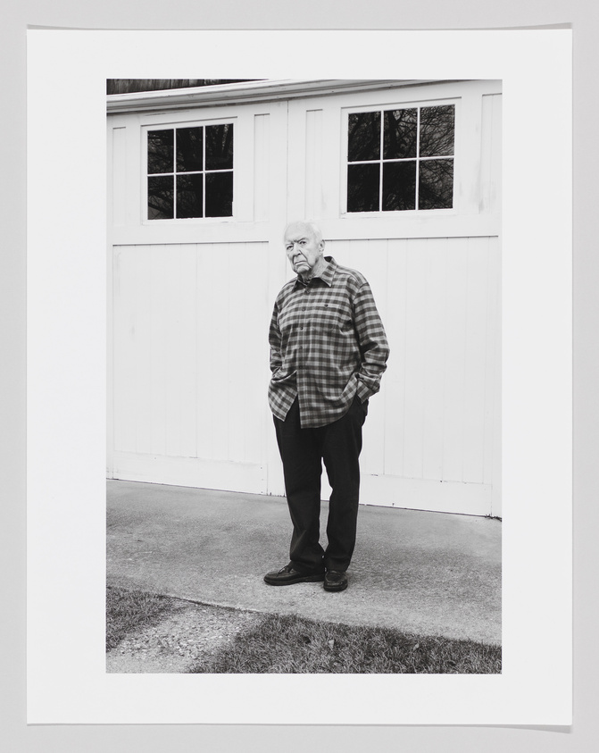 A black and white photo of Jasper Johns in a flannel standing with hands in his pockets.
