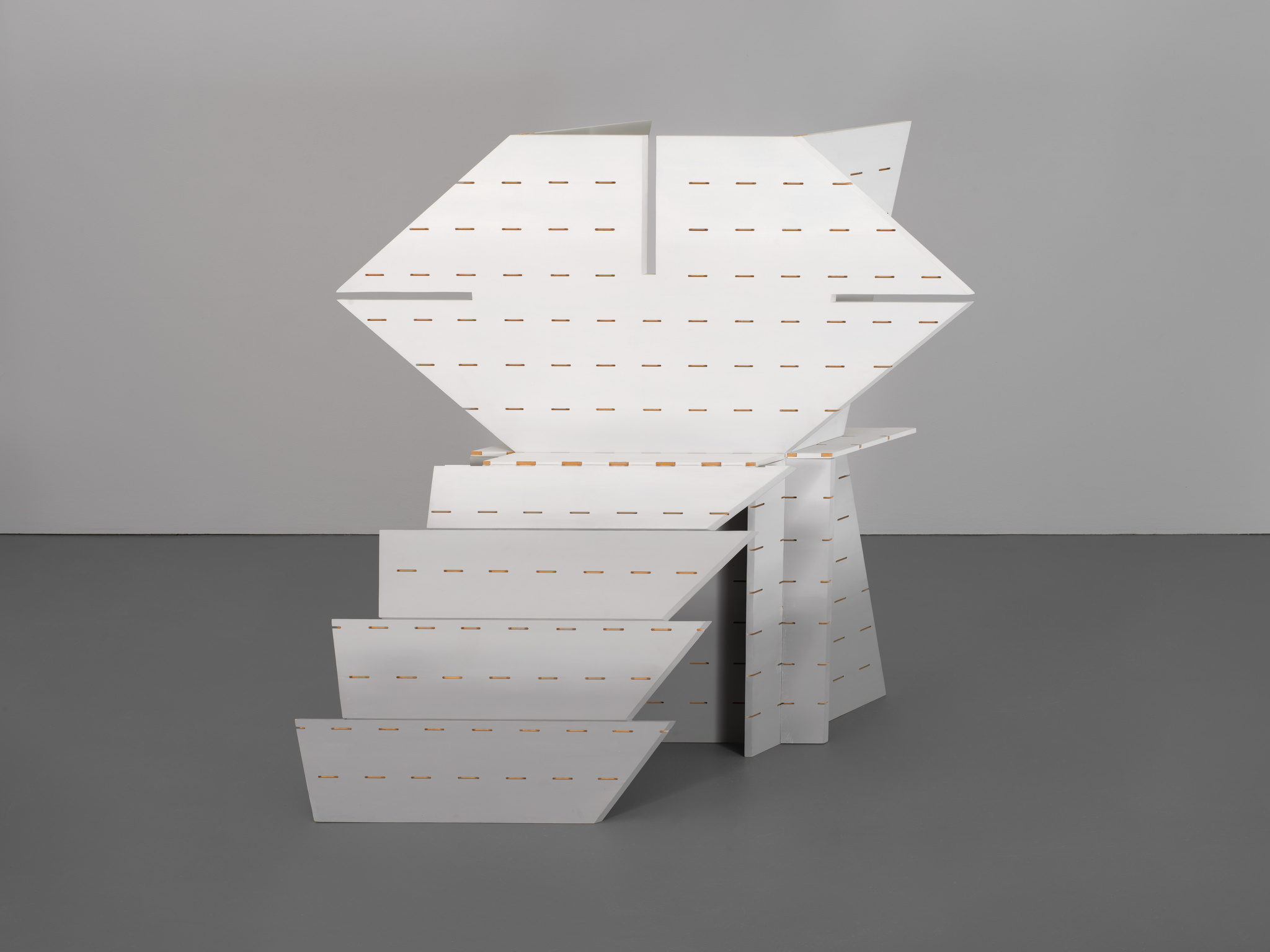 White geometric sculpture with angular shapes and horizontal slits, standing on a gray floor against a plain wall.