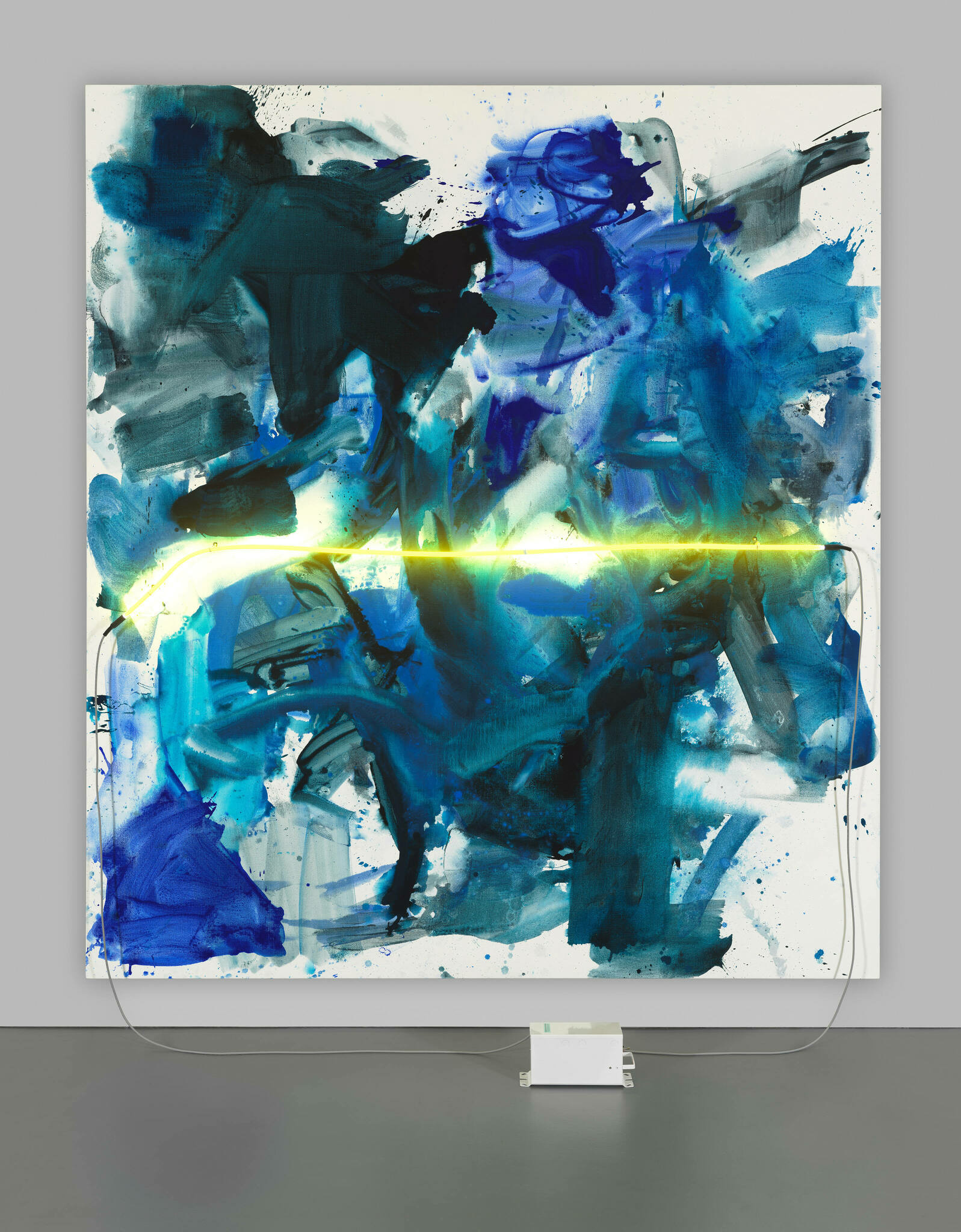 A vibrant abstract painting with various shades of blue and black splattered and smeared across the canvas, displayed in a gallery setting with a neon light strip at the bottom edge, mounted on a white wall above a dark floor. The painting is connected to a power source, suggesting the integration of light elements within the artwork.