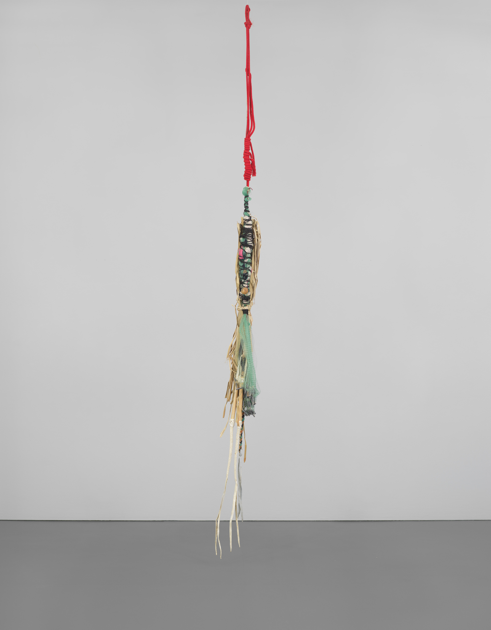 A hanging sculpture made of colorful rope, beads, fabric, and string, suspended from the ceiling by a red cord.