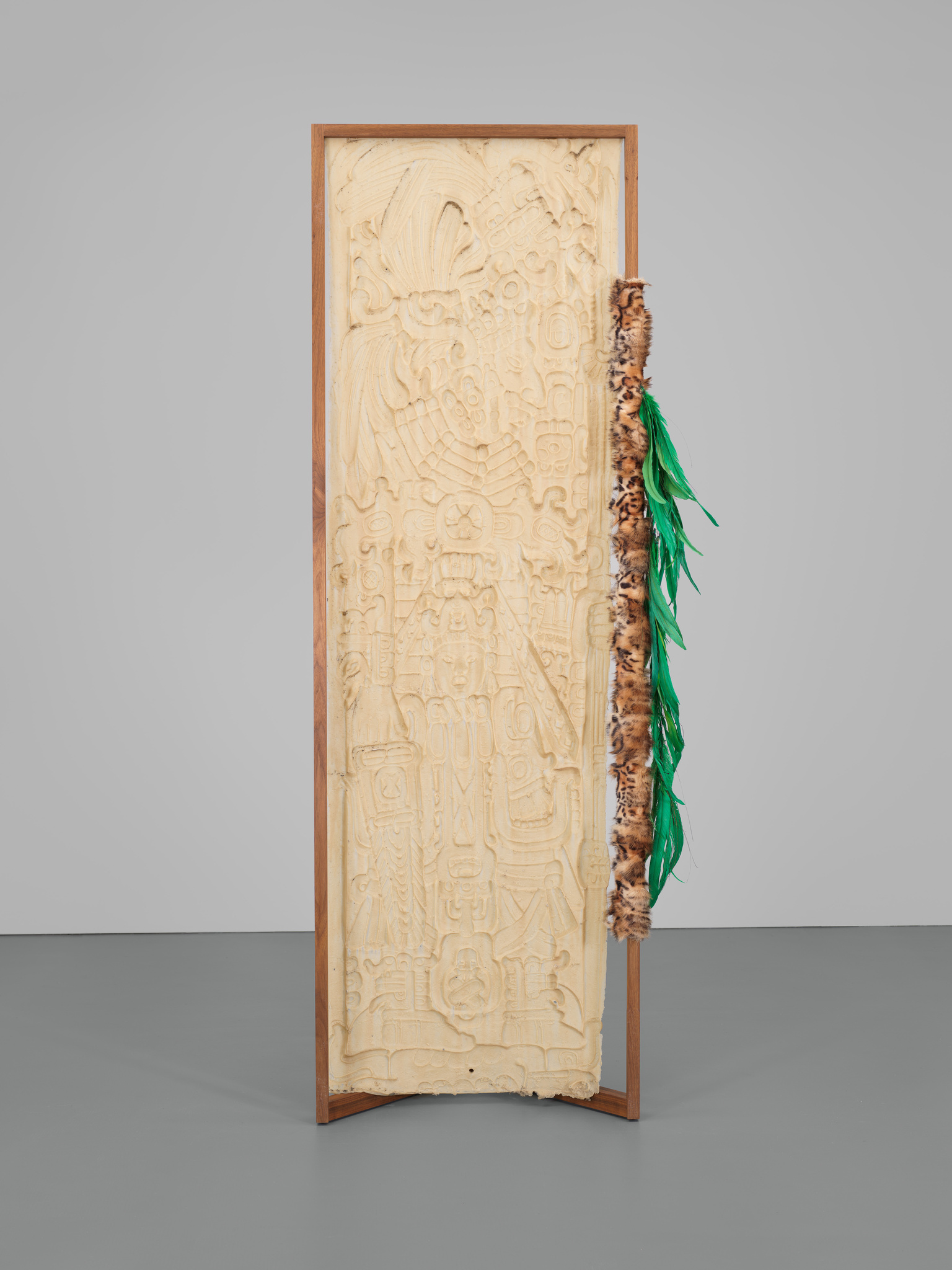 A tall, intricately carved wooden panel with a detailed relief depicting what appears to be figures and symbols, possibly of historical or cultural significance. The panel is framed with a simple wooden border and stands upright. On one side, there is a decorative element featuring green feathers and animal fur, adding a contrasting texture to the smooth carvings of the wood. The artwork is displayed against a neutral gray background.