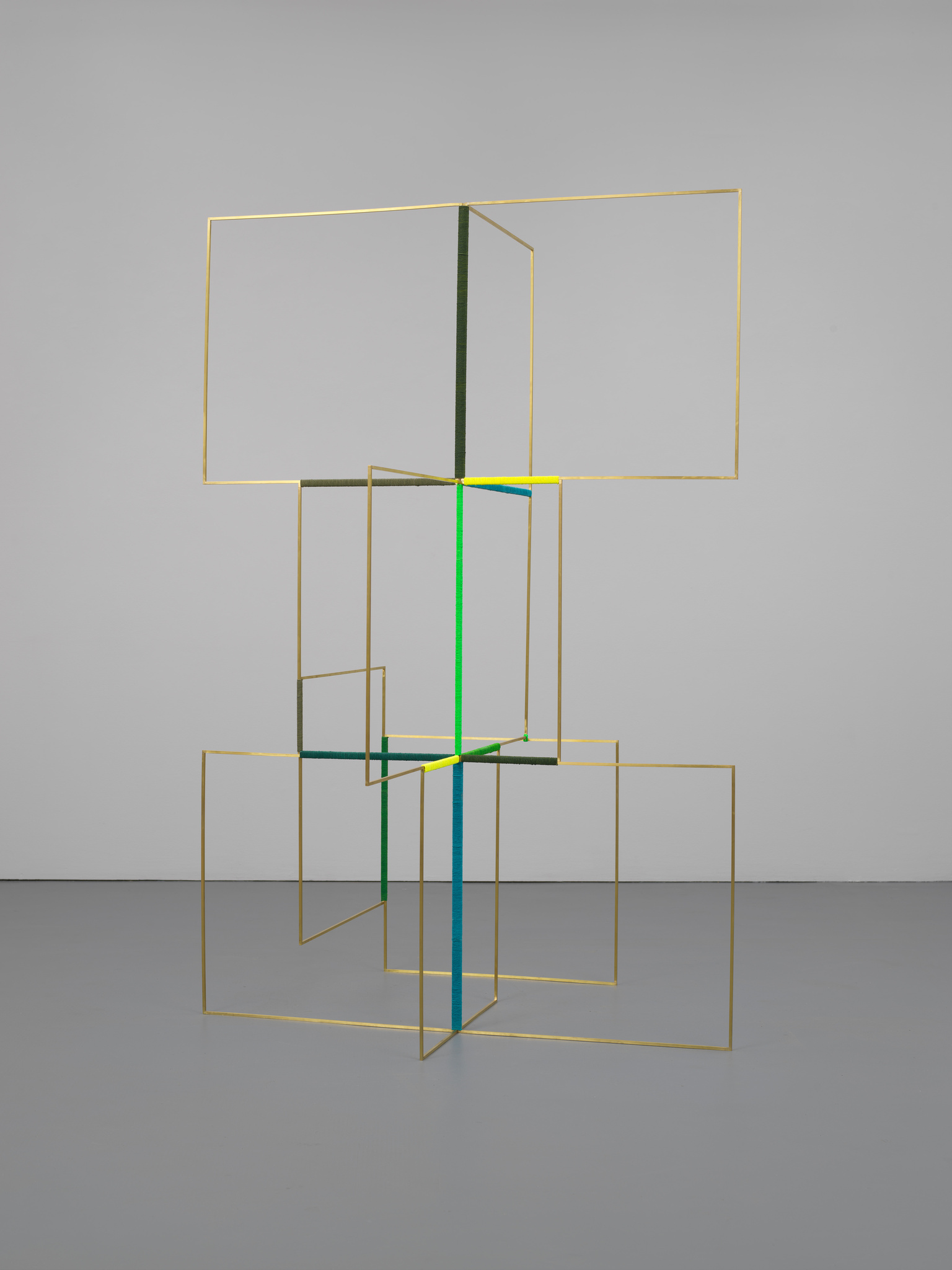 A modern art installation featuring a geometric sculpture composed of several interconnected golden frames. The frames create an abstract, three-dimensional structure with some vertical and horizontal lines highlighted in green and blue, set against a plain white gallery wall and grey floor.