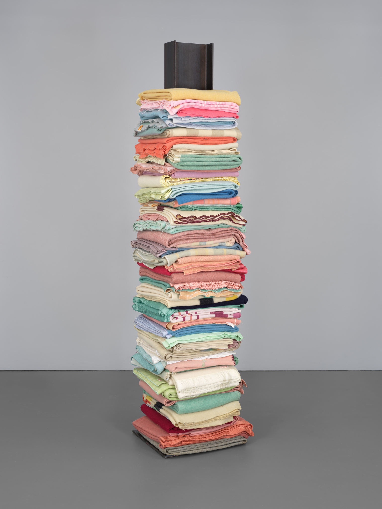Brightly-colored reclaimed wool blankets stacked in an upward pile and punctured in the center by a vertical steel I-beam