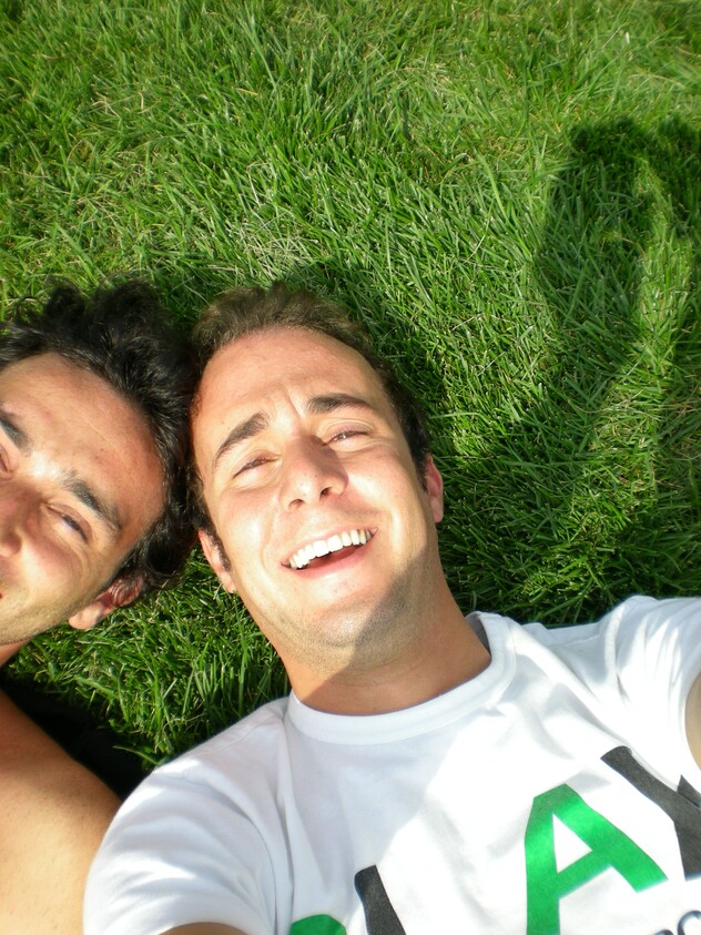 Two people lying on grass, smiling at the camera. One wears a white shirt with "PLAY" partially visible. Bright sunlight casts shadows.