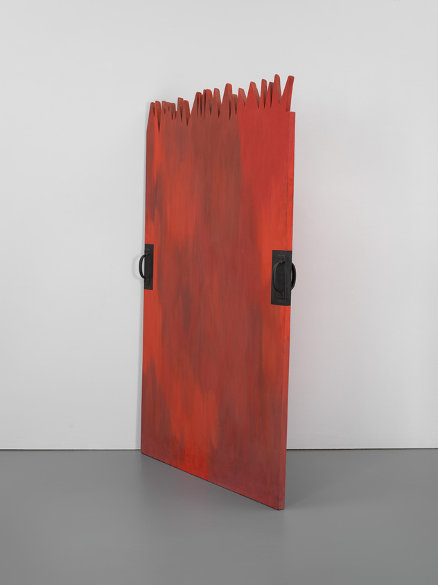 A red, rectangular wooden panel with jagged top edges and black handles on both sides, leaning against a white wall on a gray floor.