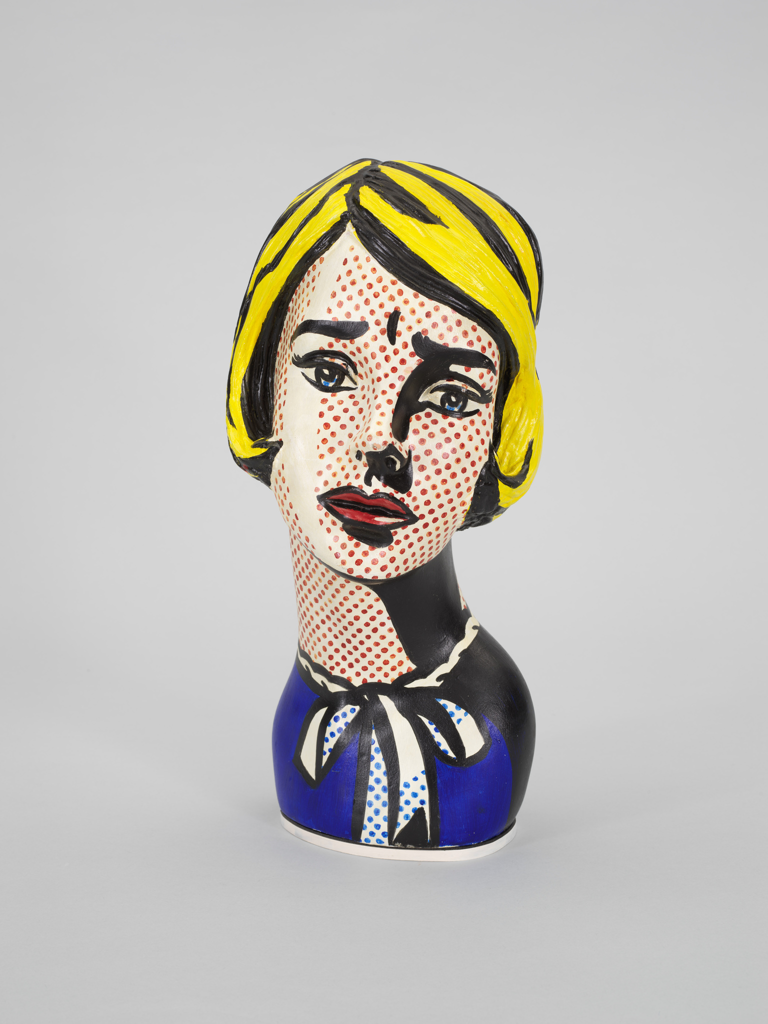 A ceramic vase painted to resemble a stylized woman's head and upper torso, with a bold pop art design featuring a yellow hairdo, red dotted skin, and a blue dress with a white collar and polka-dotted bow. The background is plain and light-colored, emphasizing the vase's vibrant colors and patterns.