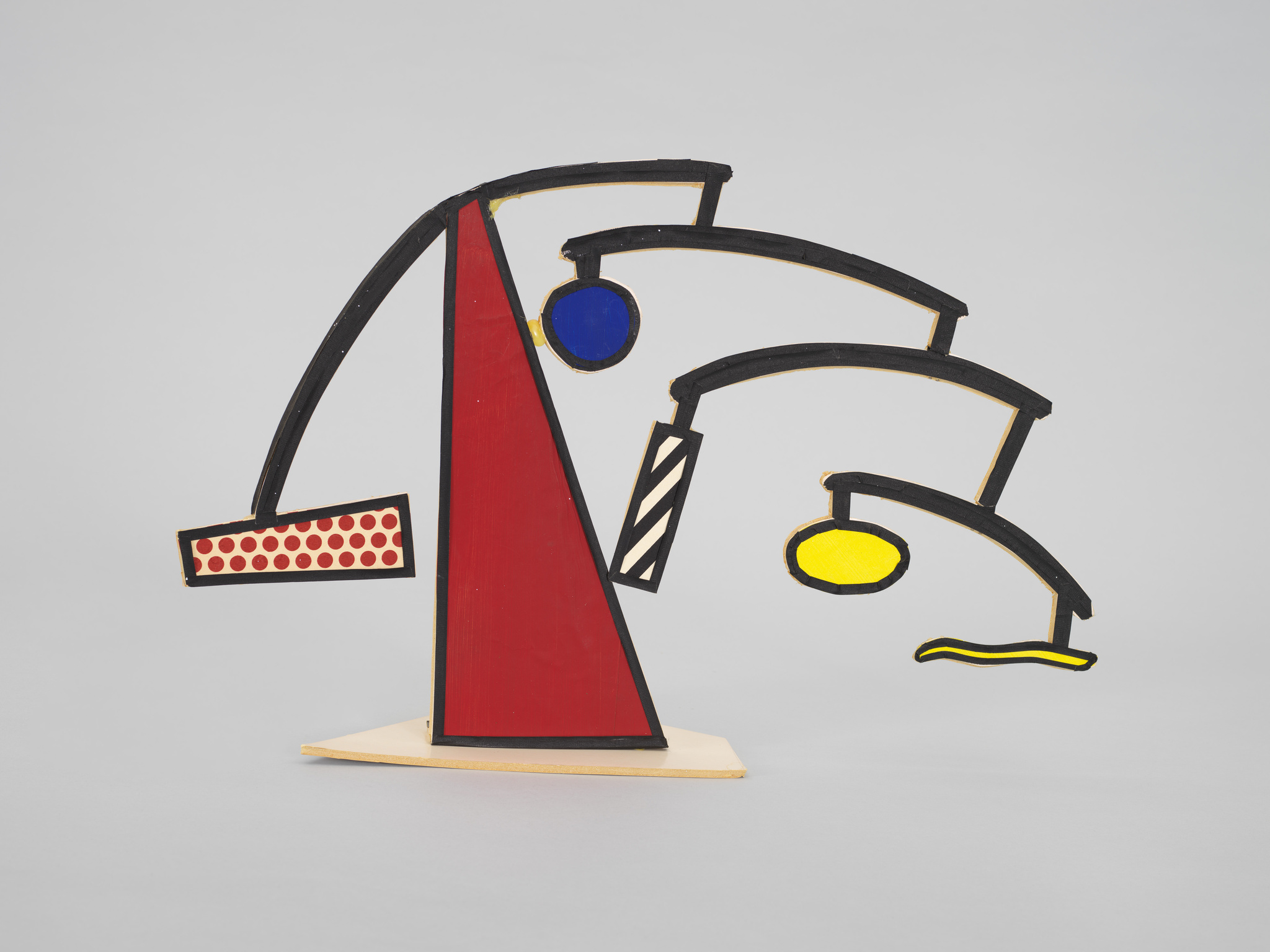 A colorful abstract sculpture featuring geometric shapes and lines in red, blue, yellow, and black, mounted on a light wooden base against a grey background.
