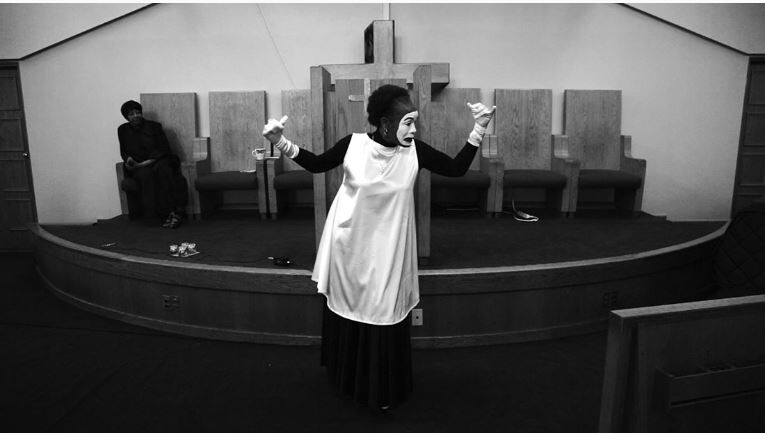 A black and white photo depicting a person dressed as a mime with expressive body language standing at the front of a church sanctuary, while another person sits contemplatively in the background to the left. The mime is wearing white gloves and a traditional mime outfit with a white face. There are scattered papers on the floor in front of the mime.