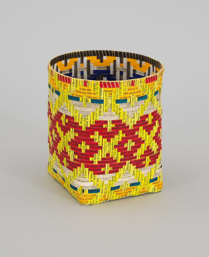 A colorful cylindrical basket woven from strips of paper printed with various words and names in different fonts and colors, predominantly yellow, red, and black, against a plain white background.