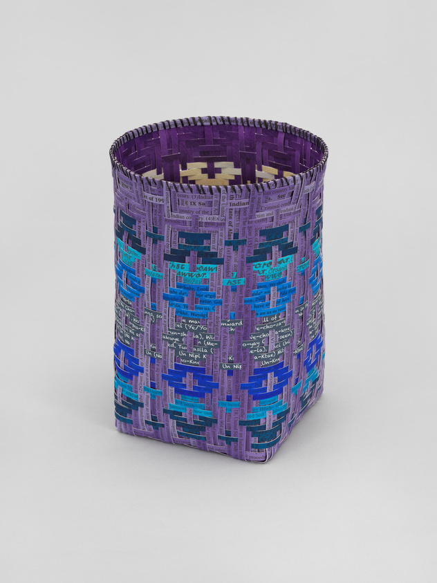 A woven basket with intricate patterns in shades of purple and blue, featuring text and numbers, suggesting it may be made from repurposed materials such as paper or fabric with printed information. The basket stands against a neutral background.