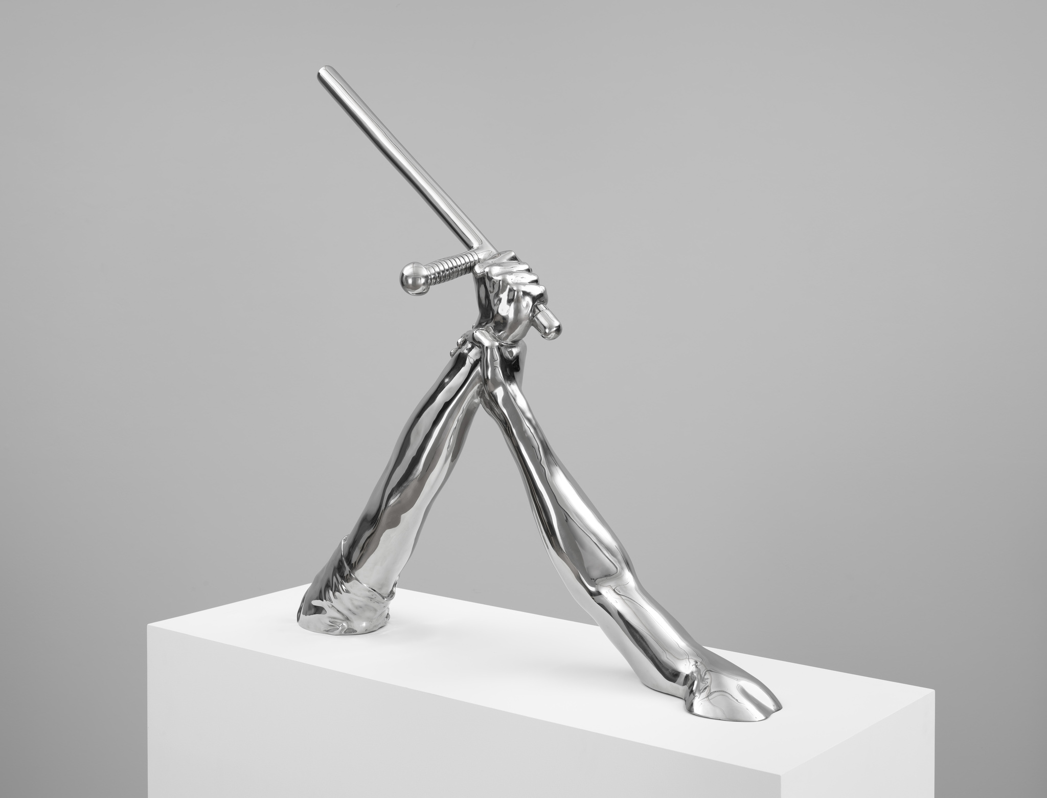A shiny, metallic sculpture of a humanoid figure with a sword, displayed on a white pedestal against a grey background.