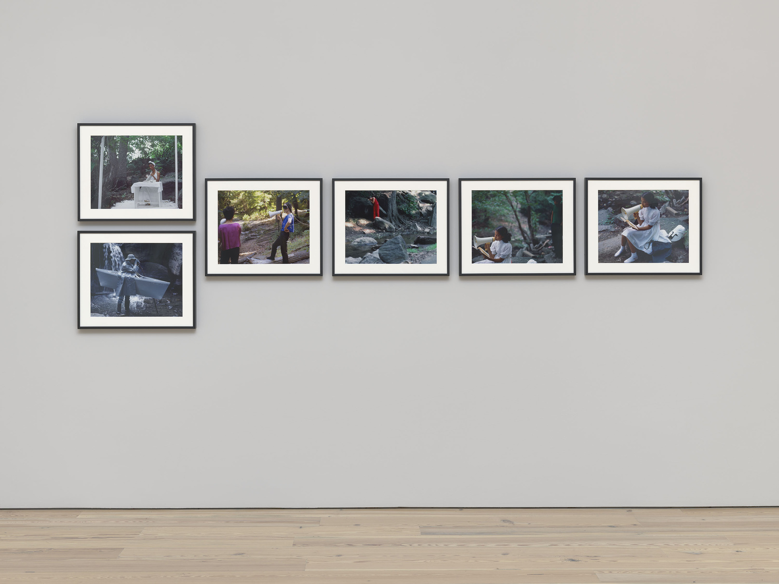 A gallery wall featuring six framed photographs of various scenes, including outdoor settings and individuals in different poses, displayed against a plain white wall above a light wooden floor.
