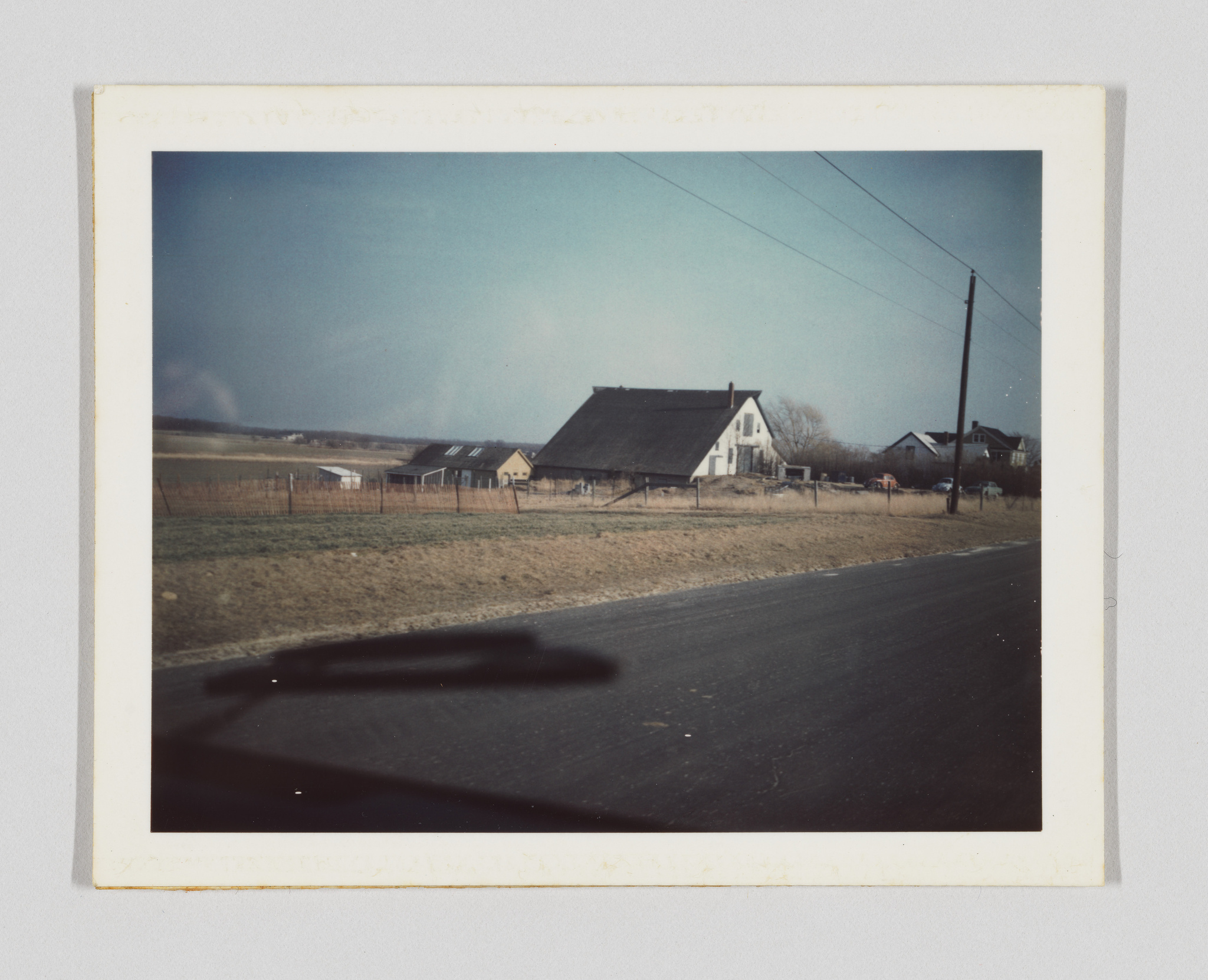 A vintage photograph depicting a rural landscape with a road in the foreground and a series of farm buildings, including a barn and a house, in the background. The sky is clear and blue, and there's a utility pole prominently visible on the right. The photo has a white border, and there's a shadow in the lower part that suggests it was taken from inside a vehicle.