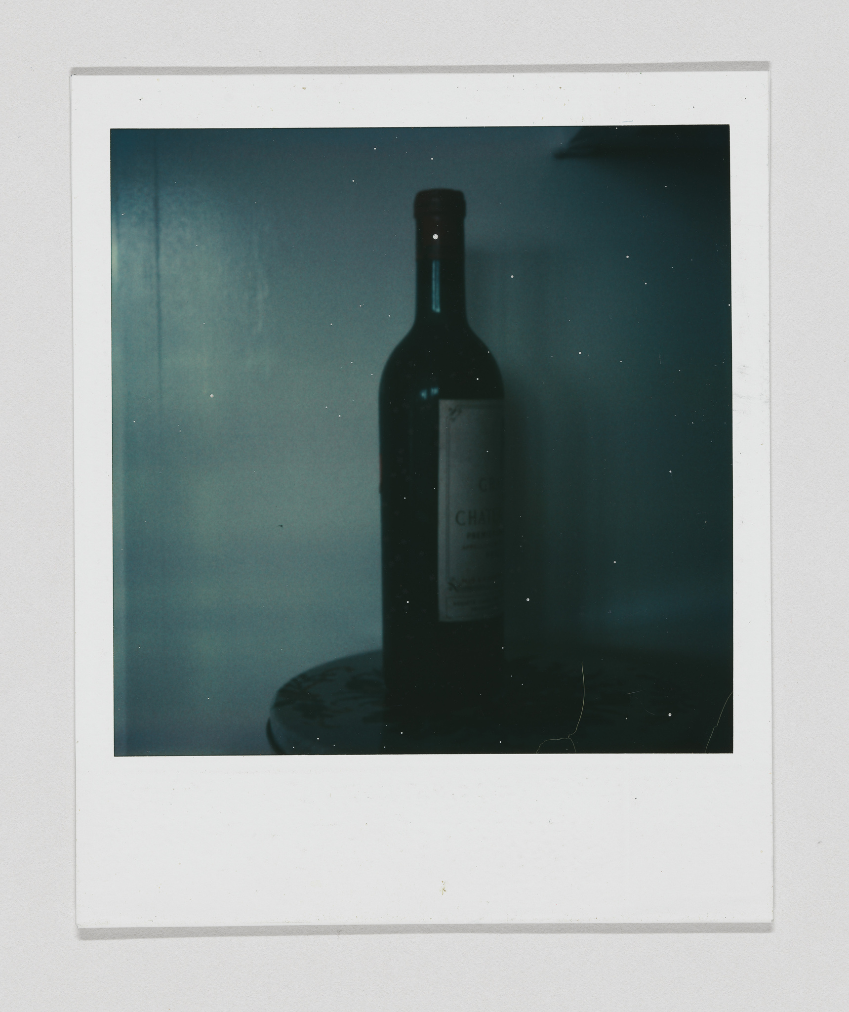 A Polaroid photo featuring a dark silhouette of a wine bottle on a plate against a dimly lit background.