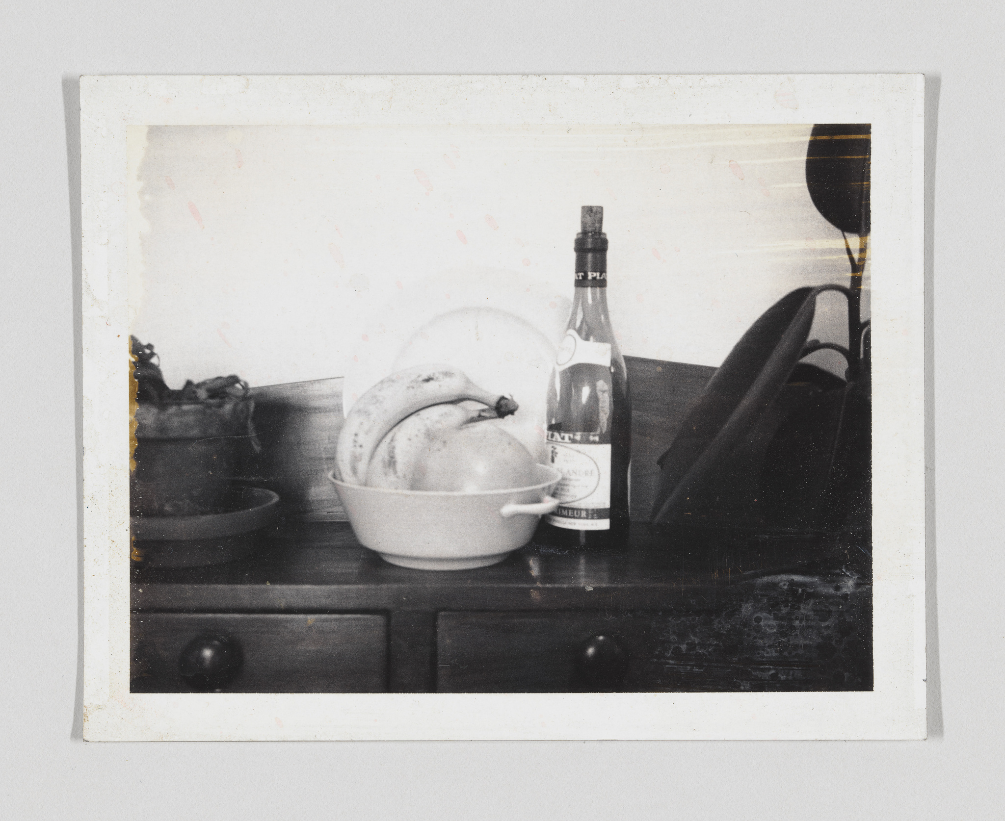 A vintage-looking photograph depicting a still life scene on a dark wooden surface. There is a bowl with bananas, a bottle of wine with a label that appears to be in French, and a dark wine glass to the right. The image has a white border with signs of wear and age, such as spots and discoloration.