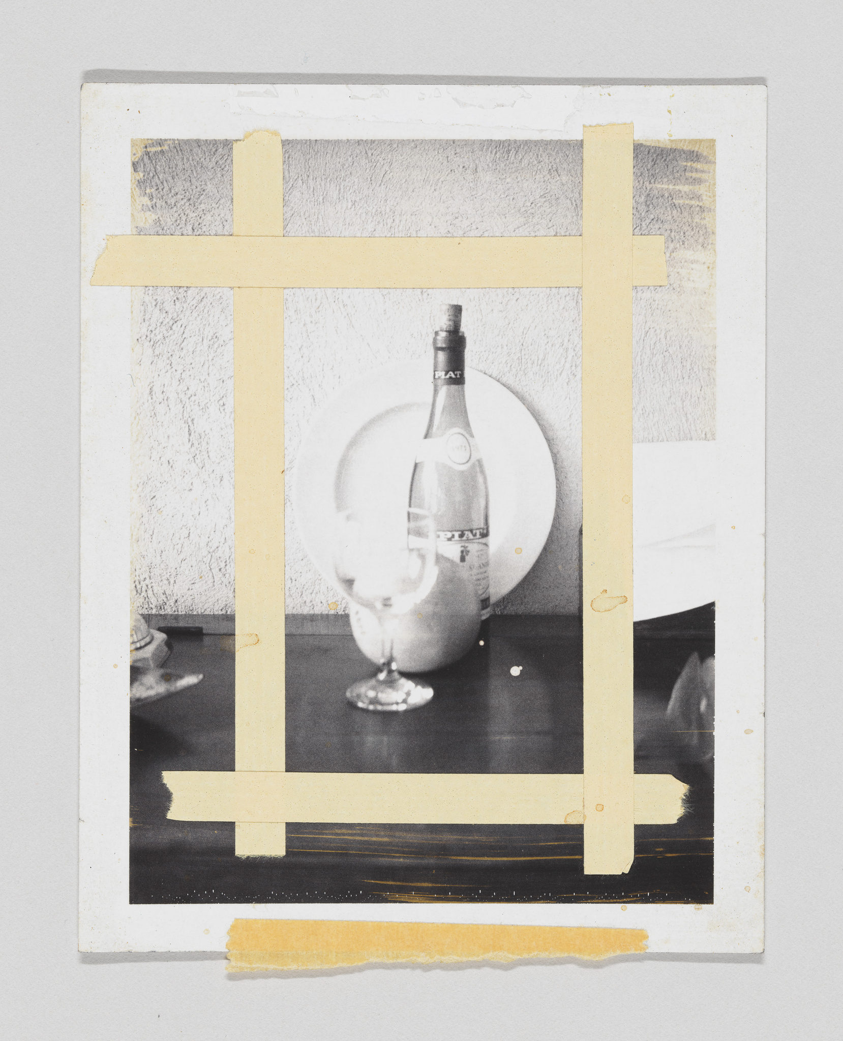 A black and white photograph of a still life scene with a bottle and a glass, partially covered by strips of yellow masking tape creating a frame within the frame. The image has a textured background and shows signs of wear around the edges.