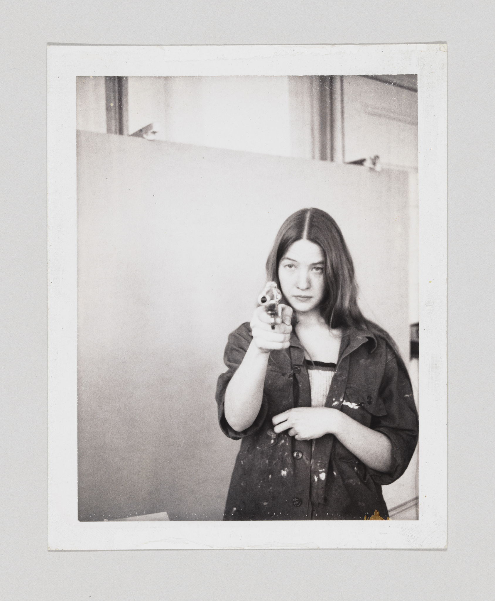 A black and white photograph of a young woman pointing a camera directly at the viewer. She has long hair parted in the middle and is wearing a dark, paint-splattered shirt over a lighter top. The photo has a white border, suggesting it is a Polaroid or instant print.