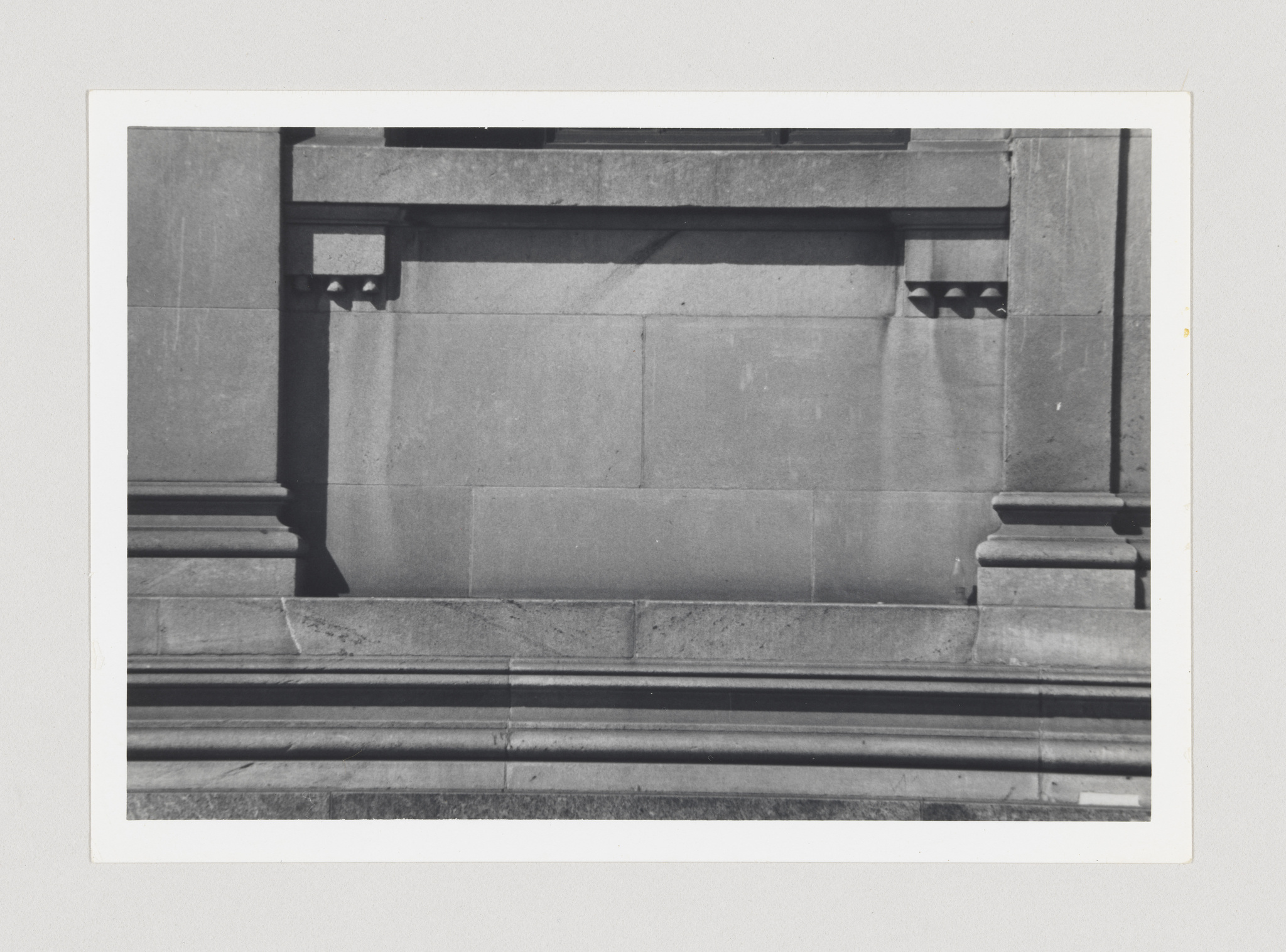 Black and white photograph of a section of a stone building with architectural details, including a shadowed alcove and ornamental brackets, framed within a white border.