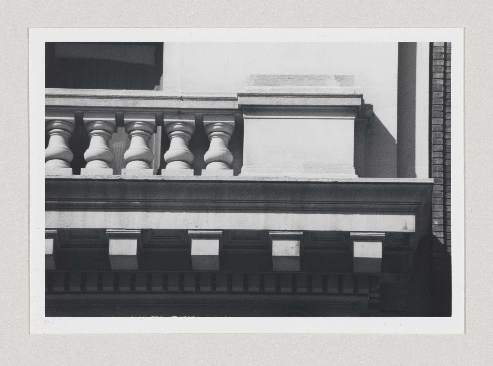 Black and white photo of architectural details showing a row of balusters on a balcony with decorative molding beneath, against a backdrop of a building's facade and a clear sky.