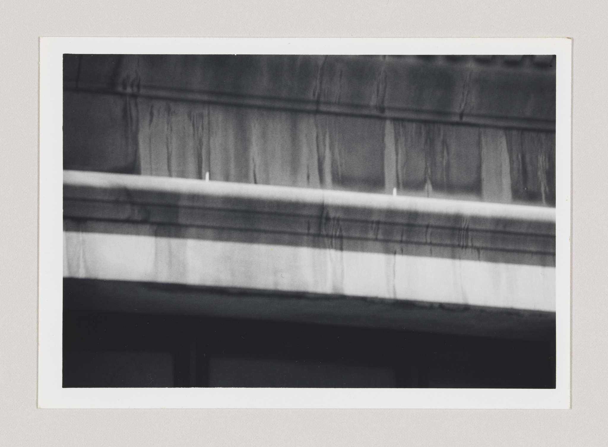 Black and white photograph of a building's concrete facade showing signs of weathering with streaks of grime or water stains running down the surface. The image is framed with a white border.
