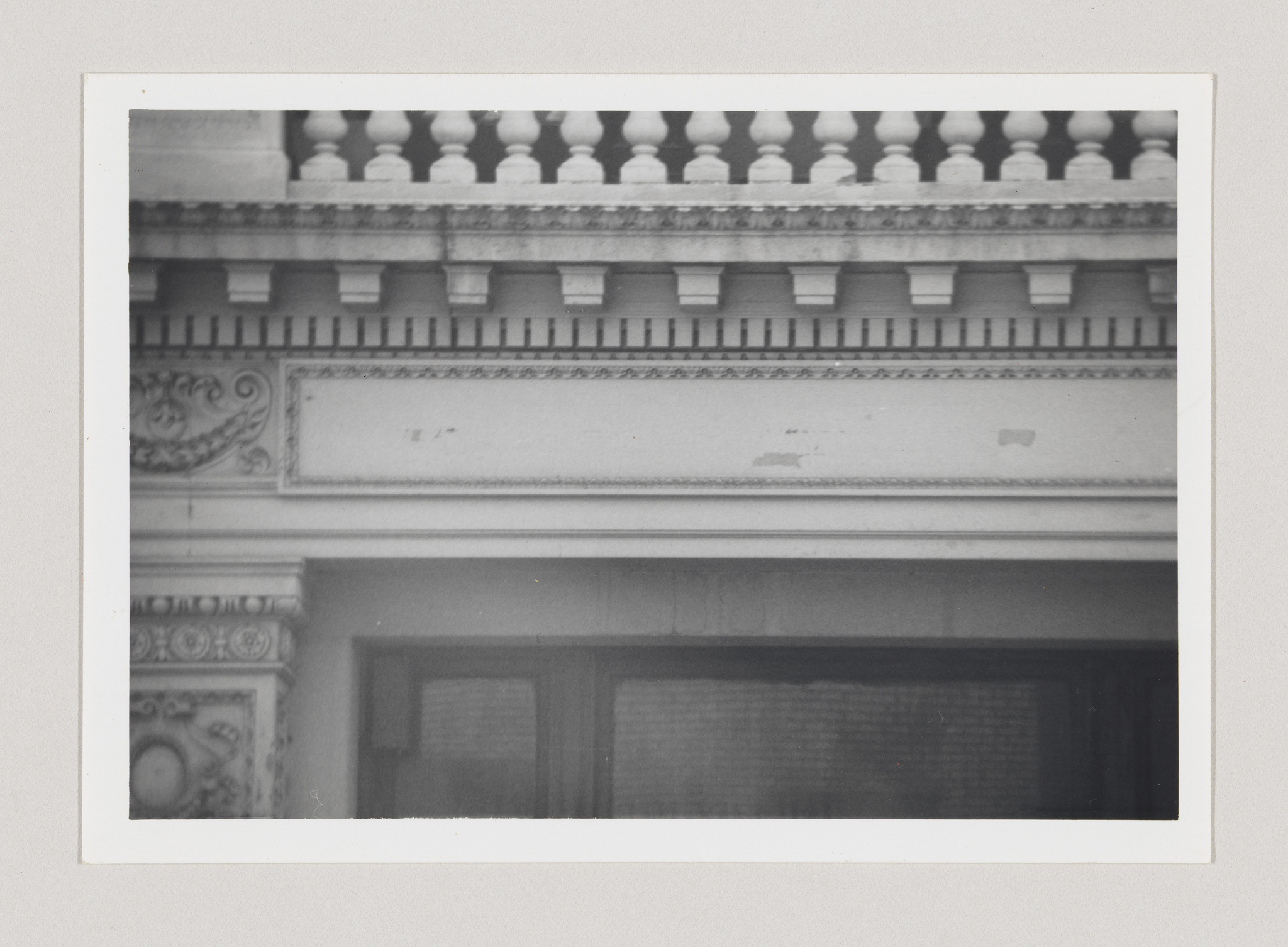 A black and white photo of architectural details showing part of a balustrade with balusters, a decorative cornice, and the upper portion of a window with a patterned curtain behind it. The image has a vintage feel, possibly taken with a film camera.