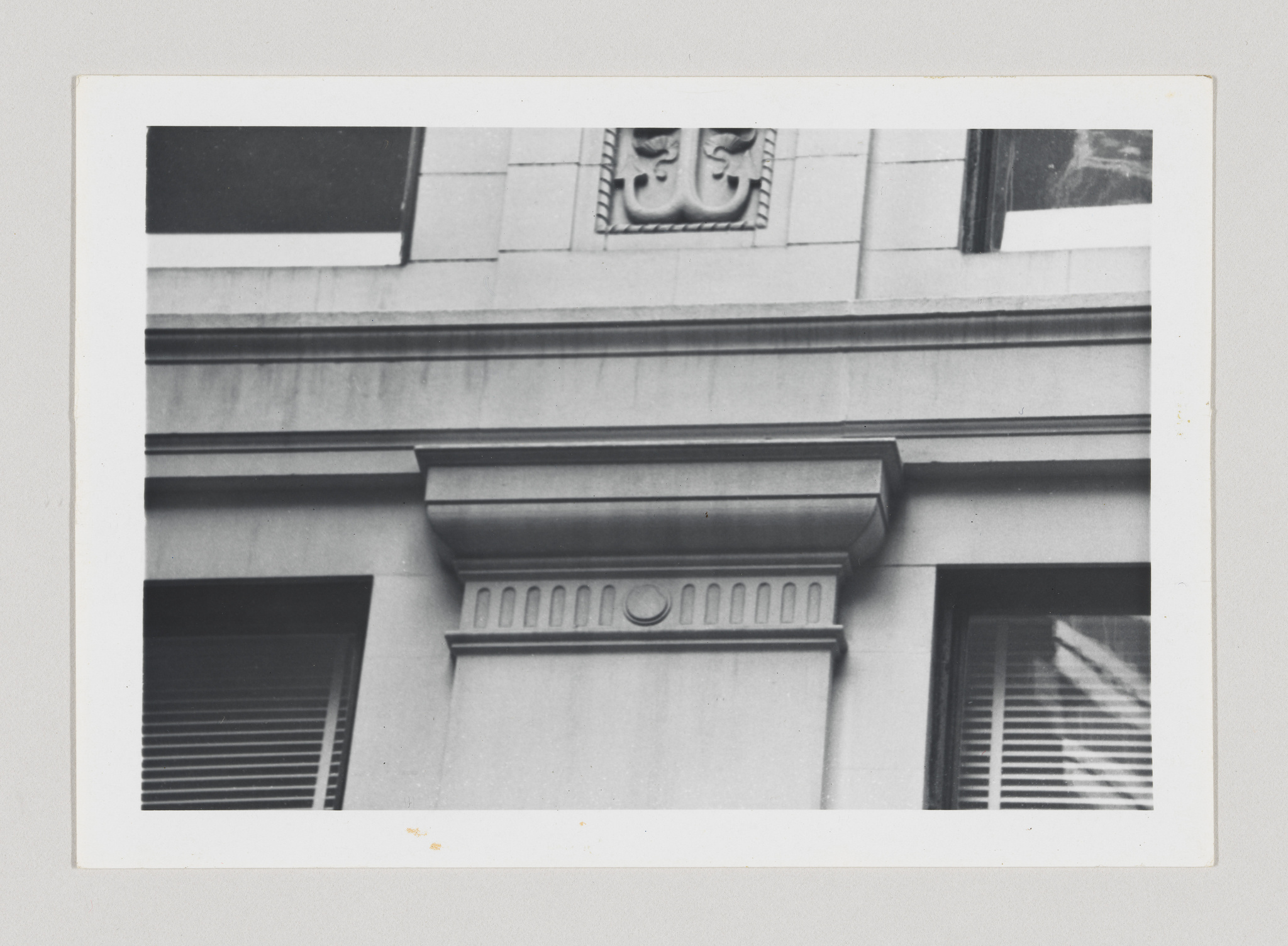 Black and white photograph of architectural details on a building facade, featuring decorative molding, a bas-relief element with a shield and foliage motif, and two windows with blinds partially drawn.