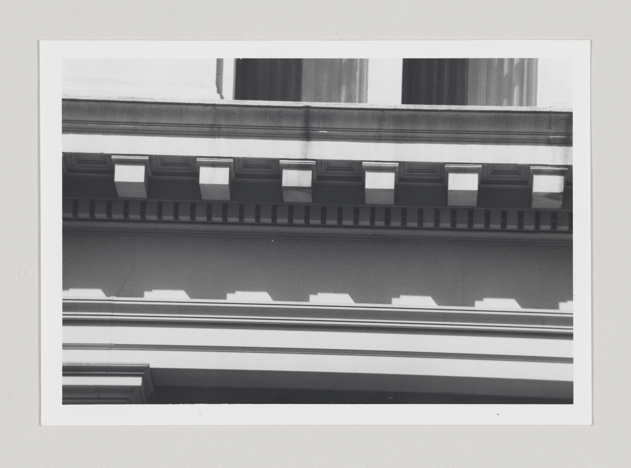 Black and white photograph showing a close-up of architectural details on a building's facade, with strong geometric patterns created by the lines and shadows of the cornice, windows, and decorative elements.