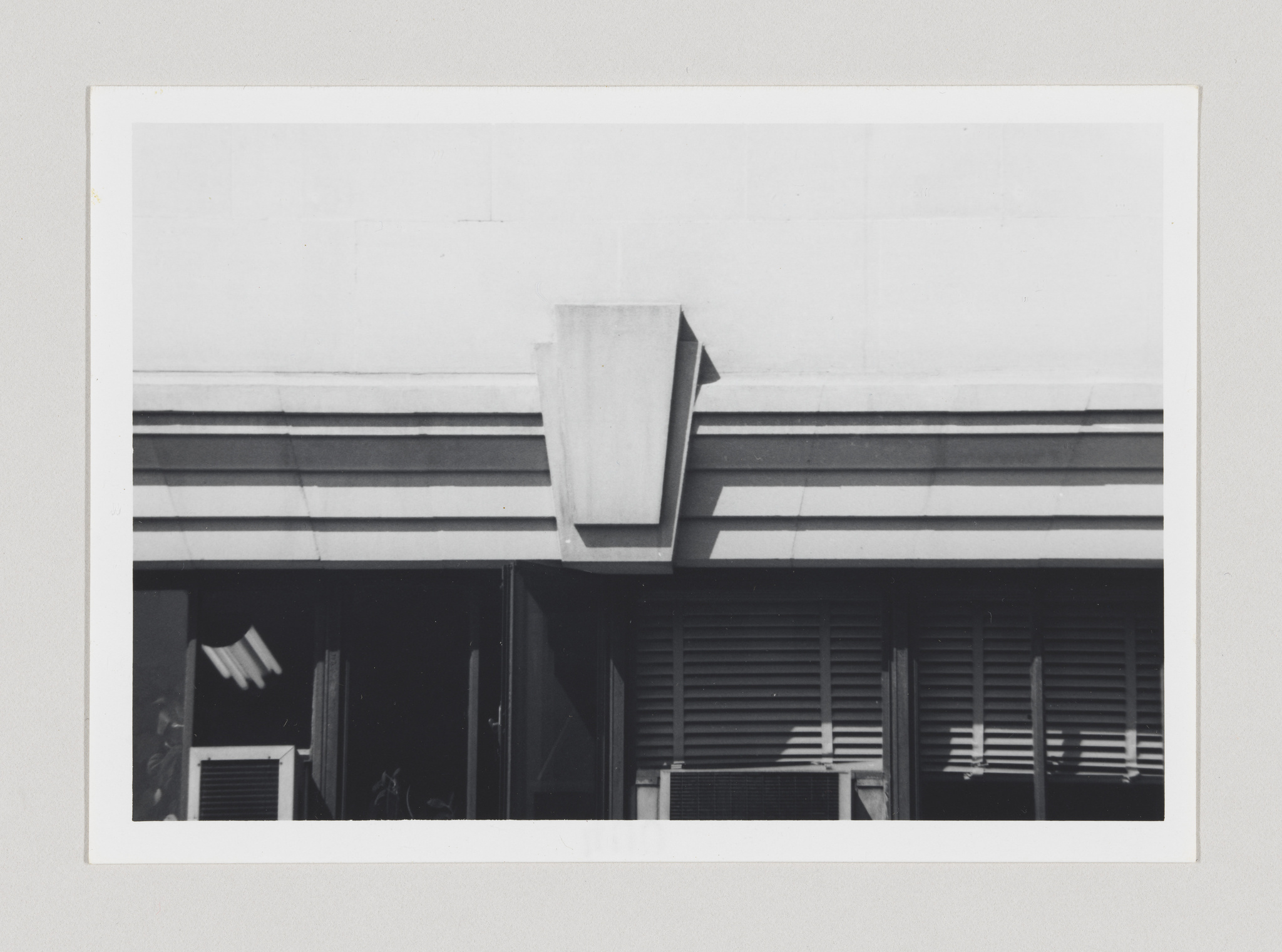 Black and white photograph of a section of a modern building facade showing geometric architectural details, including horizontal lines, a protruding rectangular structure, and shuttered windows.