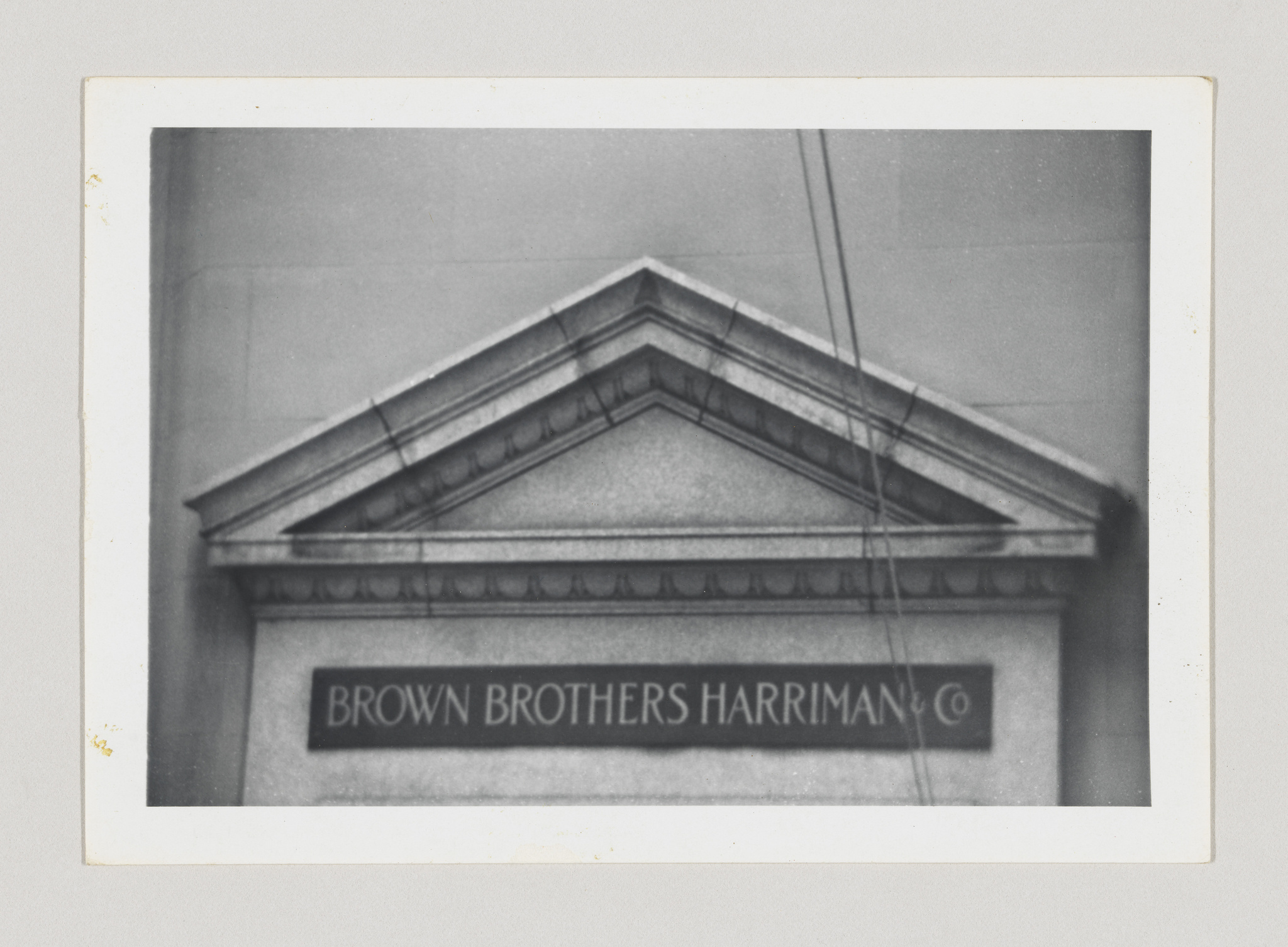 A black and white photograph of the classical pediment of a building with the name "BROWN BROTHERS HARRIMAN & CO" inscribed on it. The image has a vintage look with a white border, and there are visible creases and signs of age on the photograph.