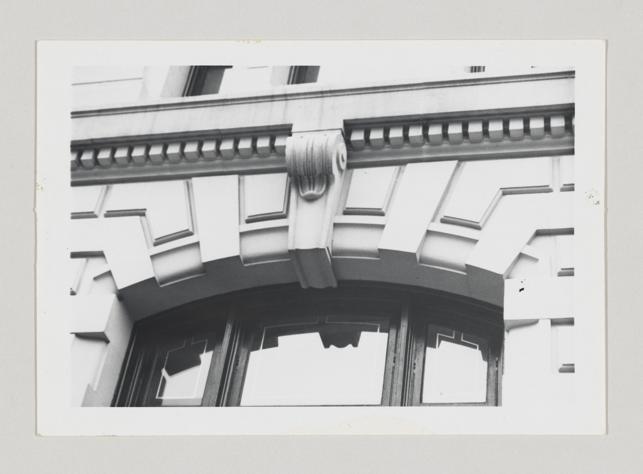 Black and white photograph of an architectural detail showing a section of a building facade with a decorative cornice and a draped sculptural element above a large arched window with broken panes.