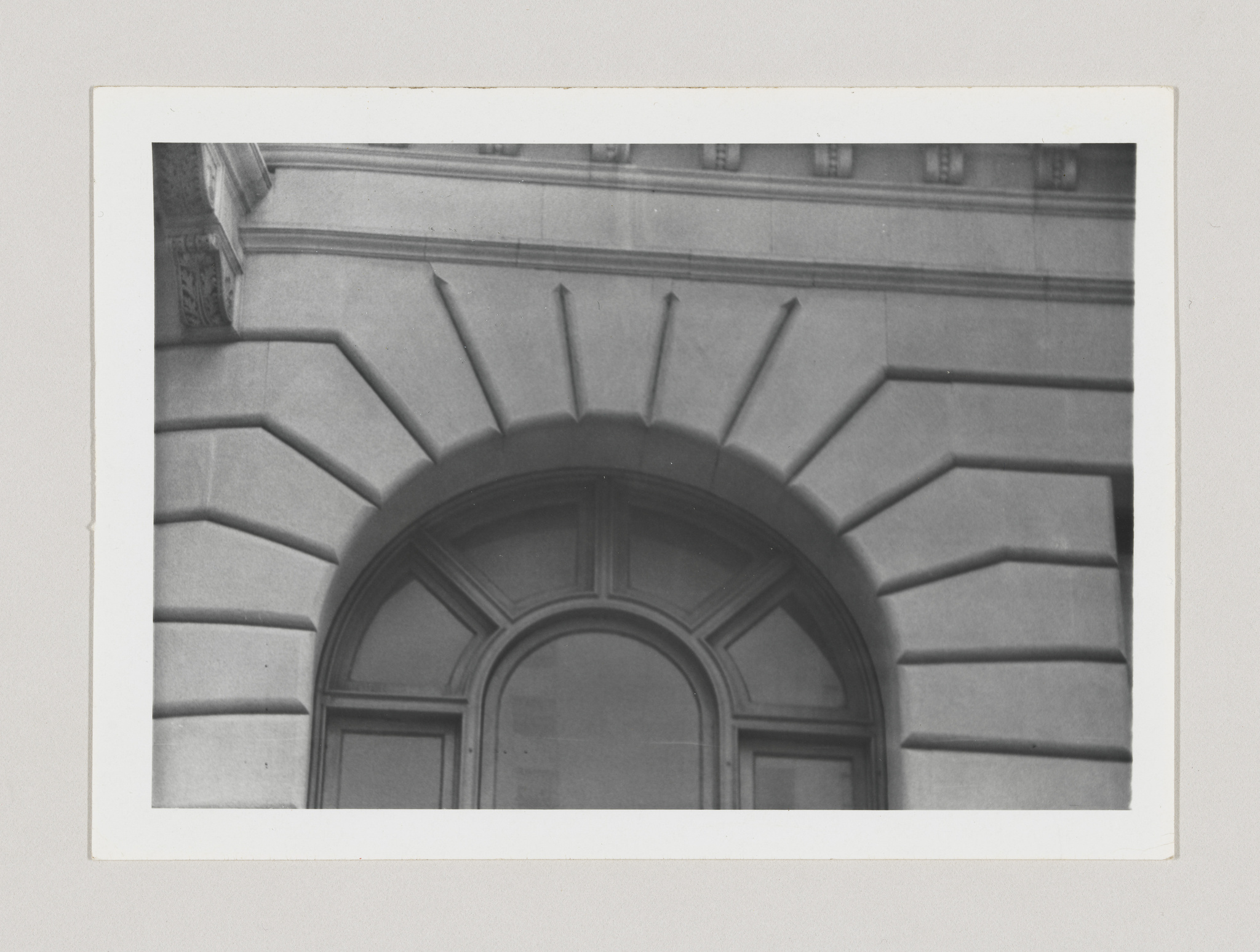 A black and white photograph of an architectural detail showing a stone archway with radiating grooves above a closed door, set within a building facade. The image has a white border, resembling a vintage photograph.