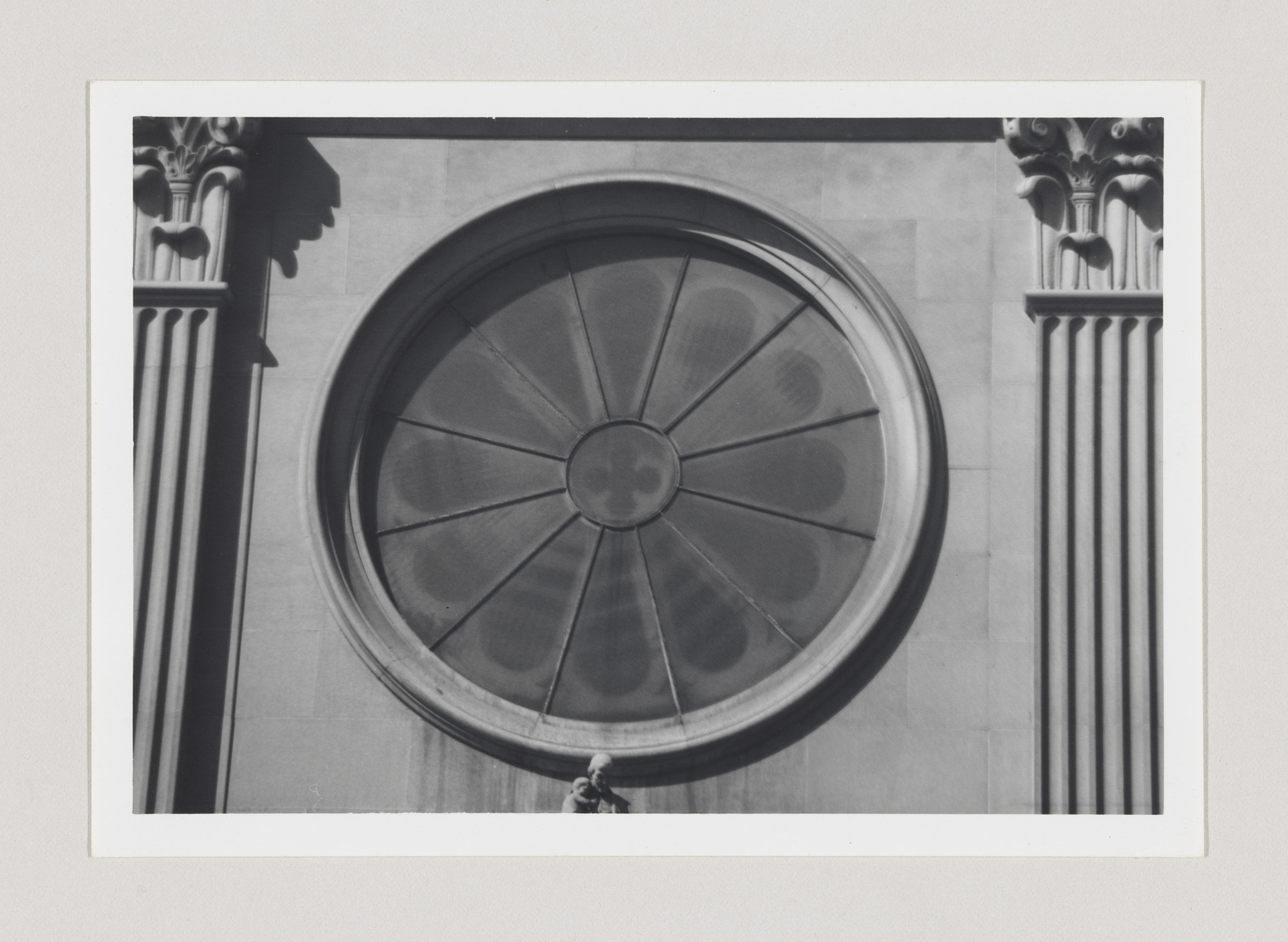A black and white photograph of a large, round window with radial divisions, set within a stone wall flanked by two classical columns with ornate capitals.