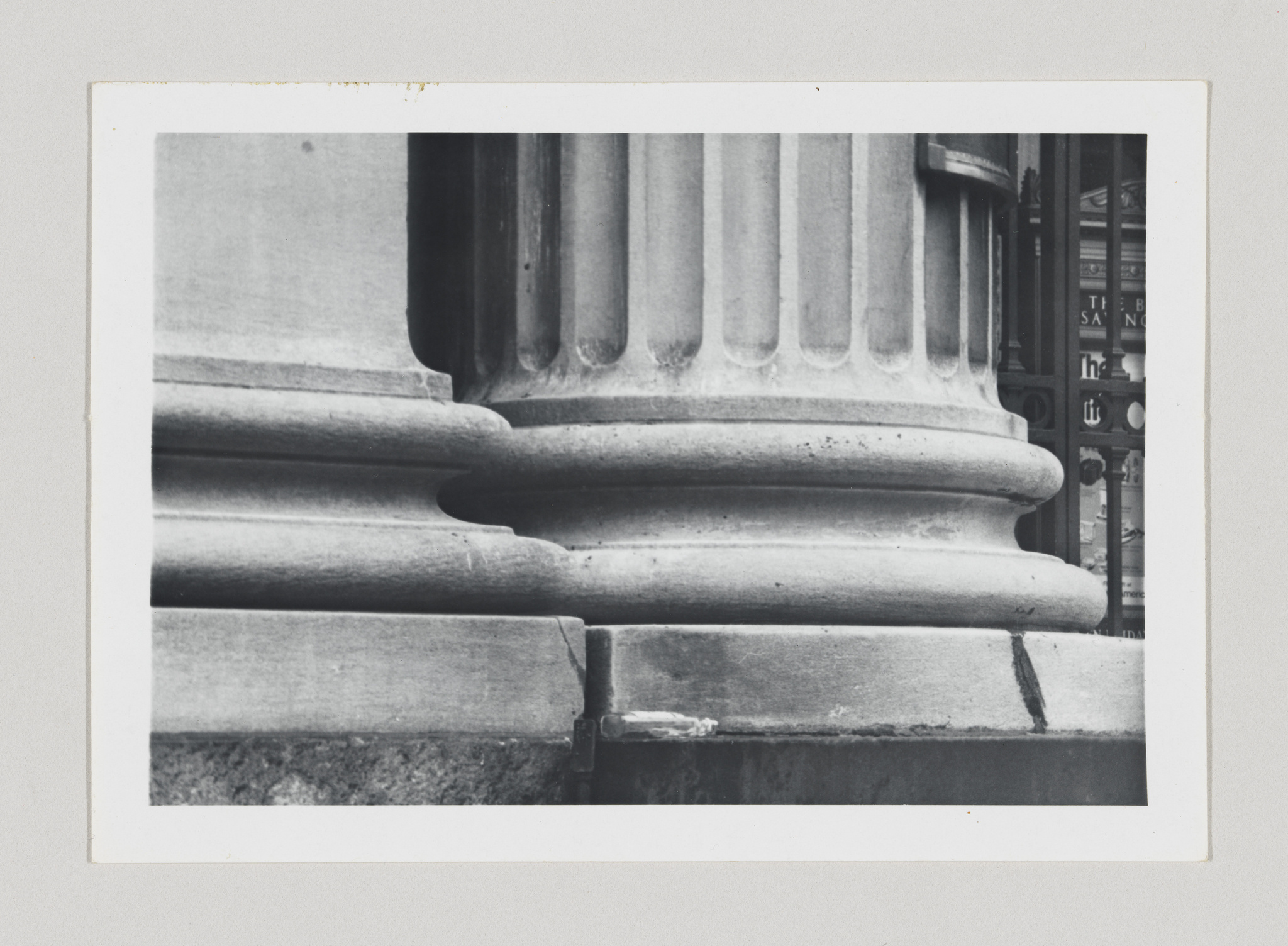A black and white photograph focusing on the base of classical architectural columns with fluted shafts, showcasing the detailed stonework and the contrast of light and shadow. In the background, partially obscured by the columns, is a glimpse of a building facade with windows and text.