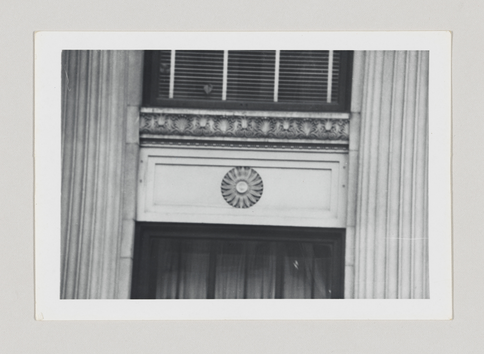 A black and white photograph of a classical architectural window detail, featuring a window with blinds, ornate molding, and a decorative rosette above the window, all framed by a pair of fluted columns. The image has a vintage appearance with a white border typical of old photographs.