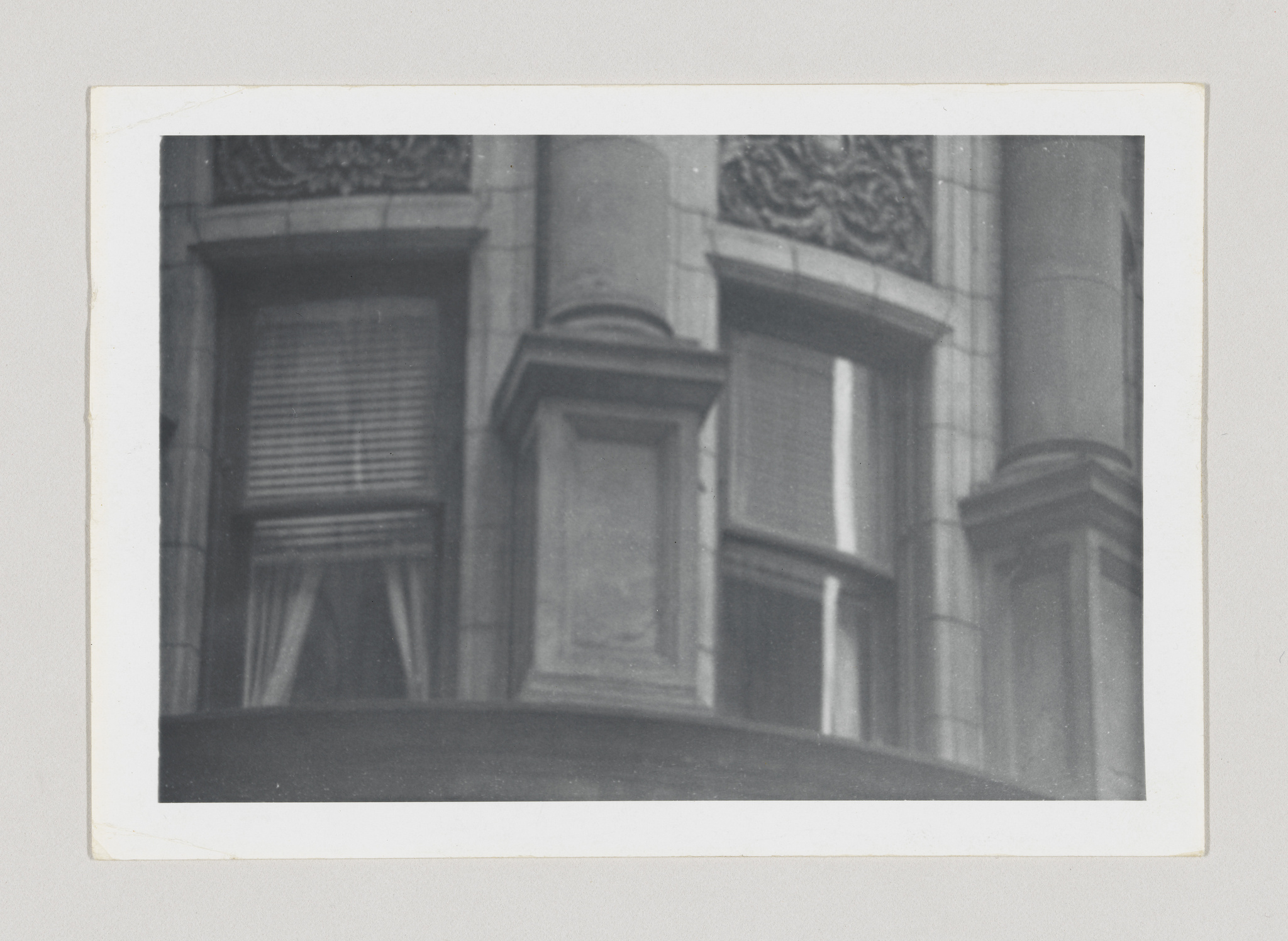 A black and white photograph of an architectural detail showing two windows with closed shutters, framed by ornate columns and decorative carvings above the windows, all presented within a slightly aged border suggesting an older photograph.