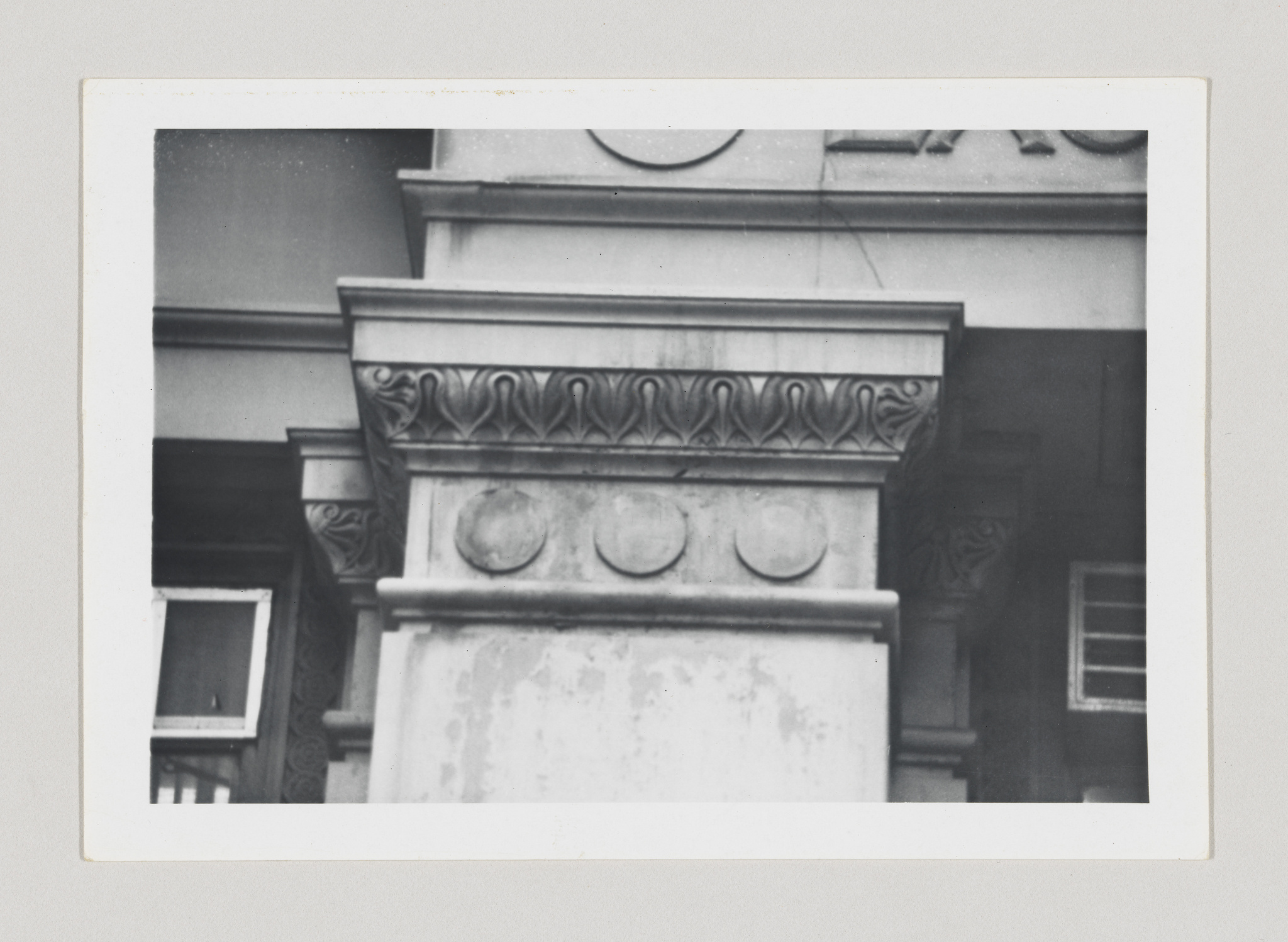 A black and white photograph of architectural details on a building, featuring classical elements such as a column with ornate capitals and a frieze with circular motifs. The image has a vintage appearance, with a white border typical of old photographs.