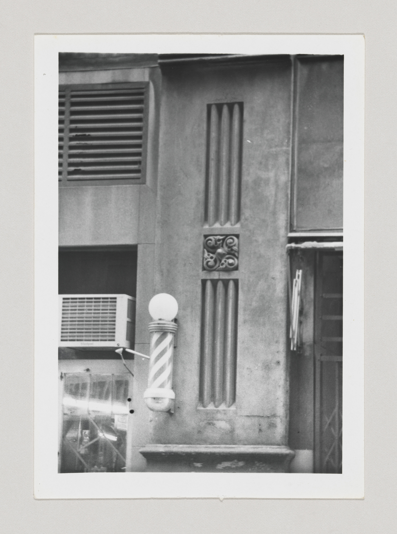 Black and white photograph of a section of a building facade showing architectural details including a window with shutters, an air conditioning unit, and a decorative column with a striped pattern and a spherical top. The image has a vintage feel, captured within a white border.
