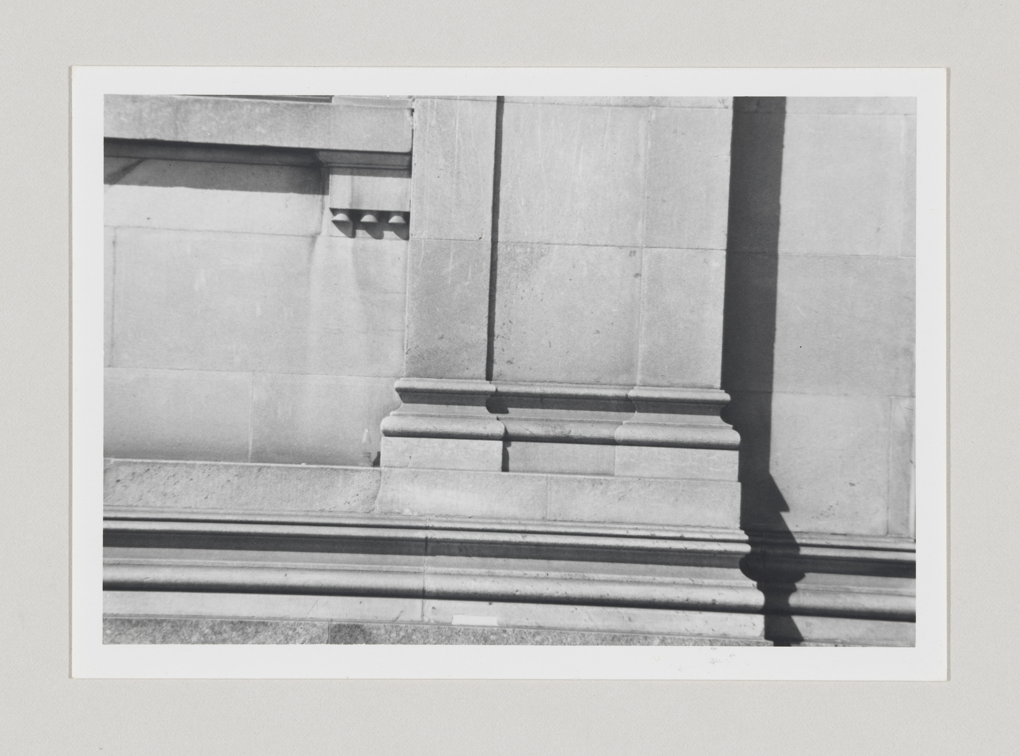 Black and white photograph of architectural details showing part of a column, its shadow, and stone wall with molding and a small decorative window grille.