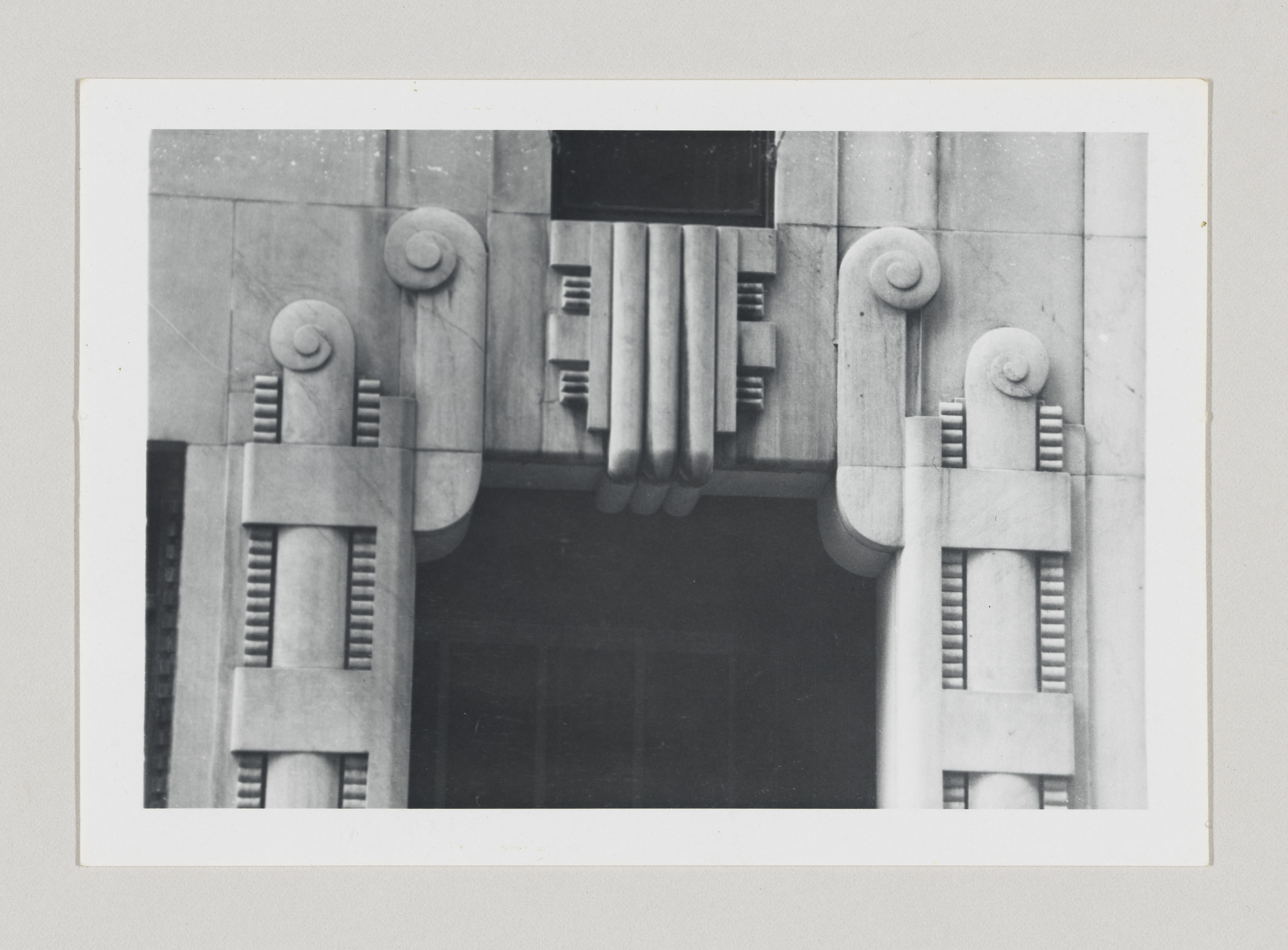 A black and white photograph of an architectural detail showing a symmetrical facade with Art Deco elements, including vertical fluting, geometric patterns, and stylized decorative scrolls on either side of a recessed dark window or opening.