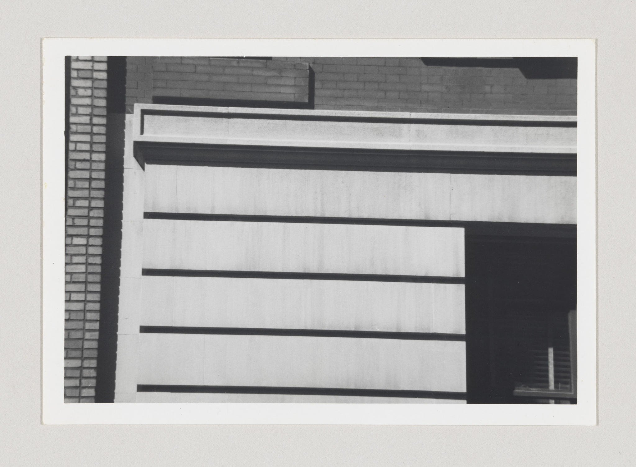 Black and white photograph of a building's corner showing contrasting geometric patterns and textures, with horizontal lines on a facade and a brick wall on the left.