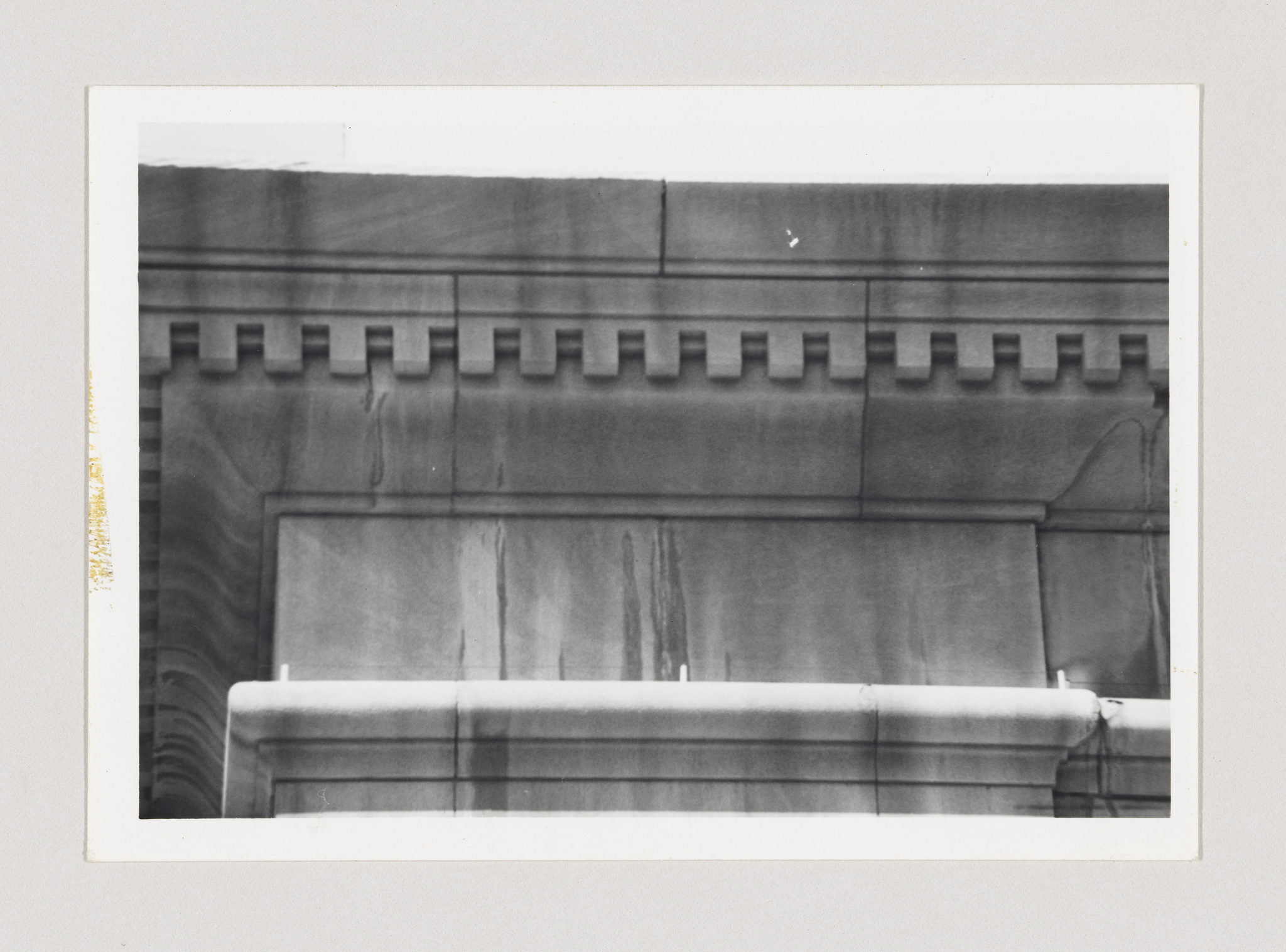 A black and white photograph of architectural details showing a section of a building with decorative molding and a horizontal pipe running across the facade. The photo appears aged with some discoloration on the edges.