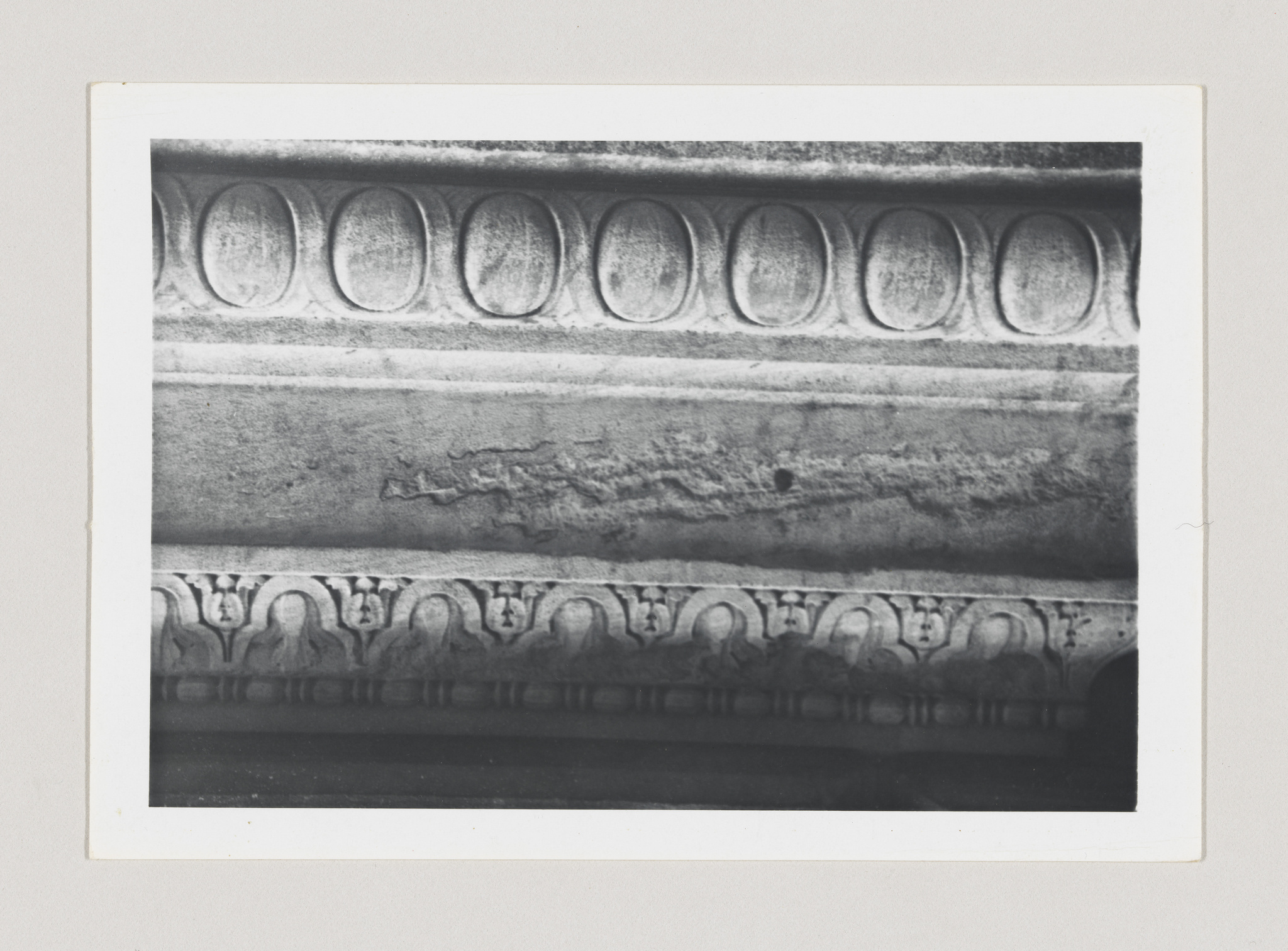A black and white photograph of an ancient stone relief with a series of oval patterns on the upper section, a damaged middle section, and a lower section featuring a row of stylized animal or human figures. The image is centered and has a white border.