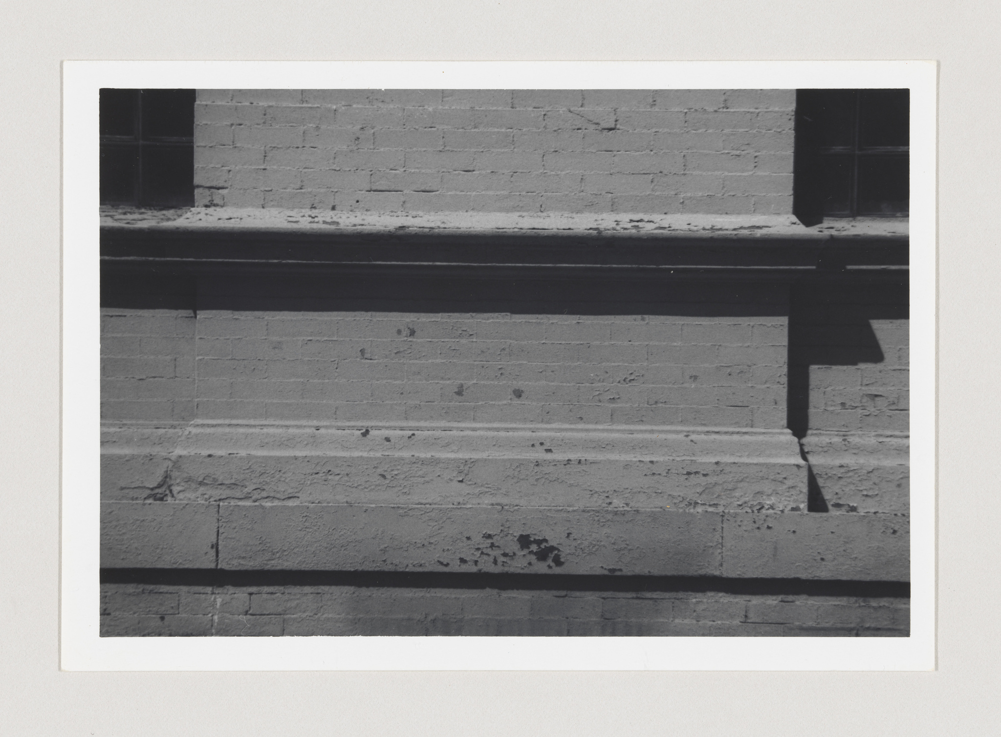 A black and white photograph of a brick wall with a single window, casting a shadow on the wall, framed with a white border.