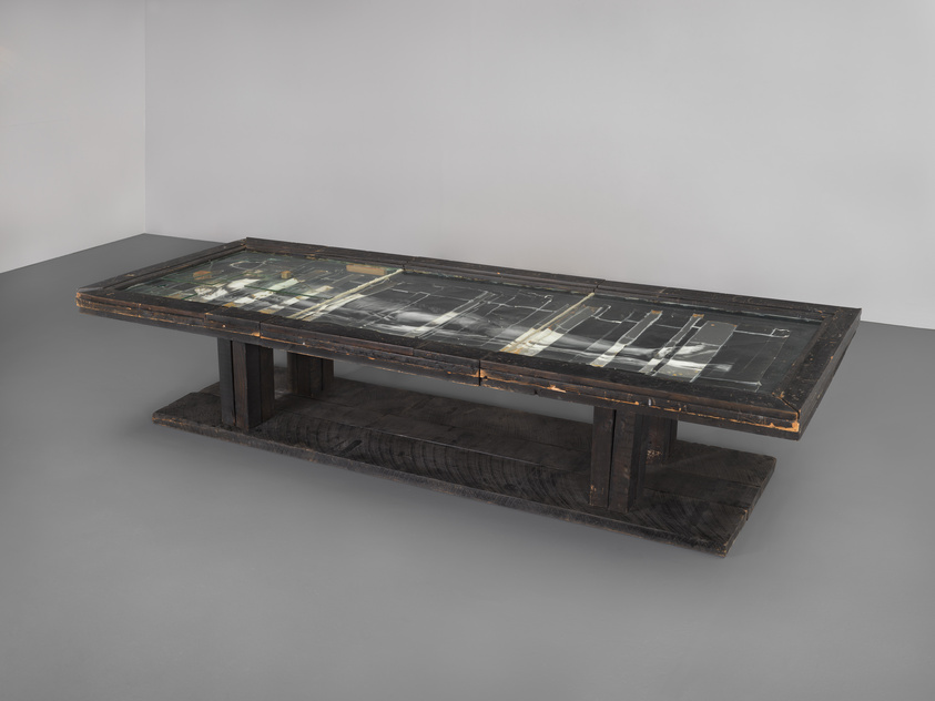 A long, dark wooden table with a glass top displaying an abstract design, set against a plain gray background.
