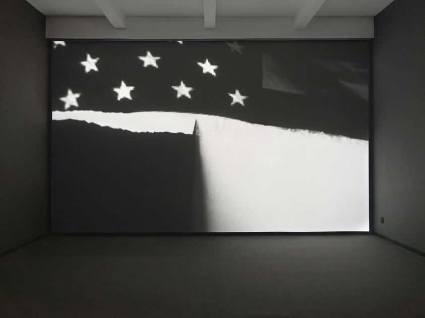 A large, dark room with a projection on the wall showing a black and white image of a starry sky, with the silhouette of a triangular shape pointing upwards at the center of the projection, creating a stark contrast with the bright background.