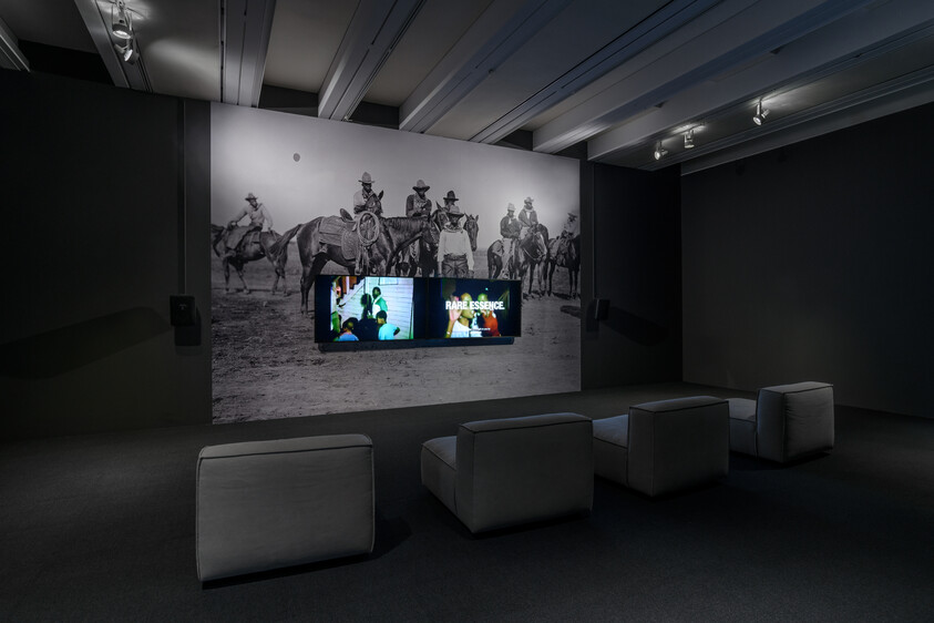 A dark room with a large black-and-white mural of cowboys and a screen showing "Rare Essence."